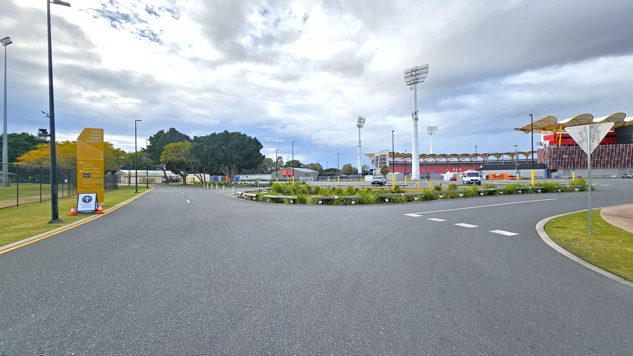 A wide road curves to the right, bordered by a grassy area with a few small plants. On the left, a tall yellow sign reads "Gold Coast Sports Precinct" with additional information below. Several tall floodlights are visible in the background, along with a large stadium structure featuring colourful vertical panels. A few vehicles are parked near the stadium, and trees are scattered throughout the area. The sky is overcast. A wide road curves to the right, bordered by a grassy area with a few small plants. On the left, a tall yellow sign reads "Gold Coast Sports Precinct" with additional information below. Several tall floodlights are visible in the background, along with a large stadium structure featuring colourful vertical panels. A few vehicles are parked near the stadium, and trees are scattered throughout the area. The sky is overcast.