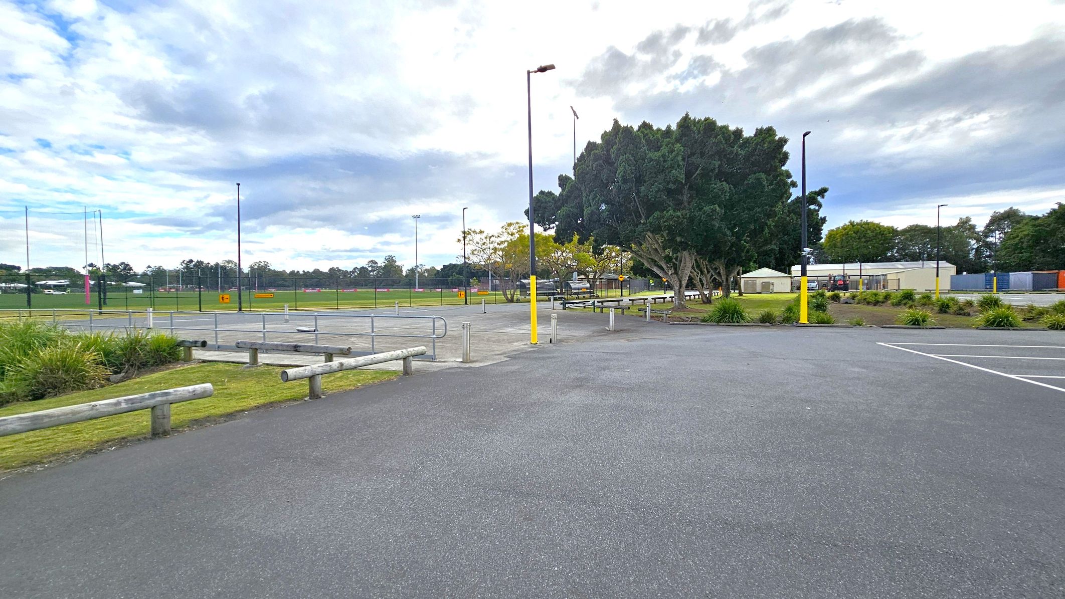 A paved area with parking spaces in the foreground, marked by white lines. Wooden barriers and small patches of grass with shrubs are on the left. In the middle, there are several tall lamp posts with yellow bases. The background features a sports field enclosed by a fence, with goalposts visible. To the right, there are trees and a few small buildings, possibly storage units or sheds. The sky is overcast with clouds. A paved area with parking spaces in the foreground, marked by white lines. Wooden barriers and small patches of grass with shrubs are on the left. In the middle, there are several tall lamp posts with yellow bases. The background features a sports field enclosed by a fence, with goalposts visible. To the right, there are trees and a few small buildings, possibly storage units or sheds. The sky is overcast with clouds.
