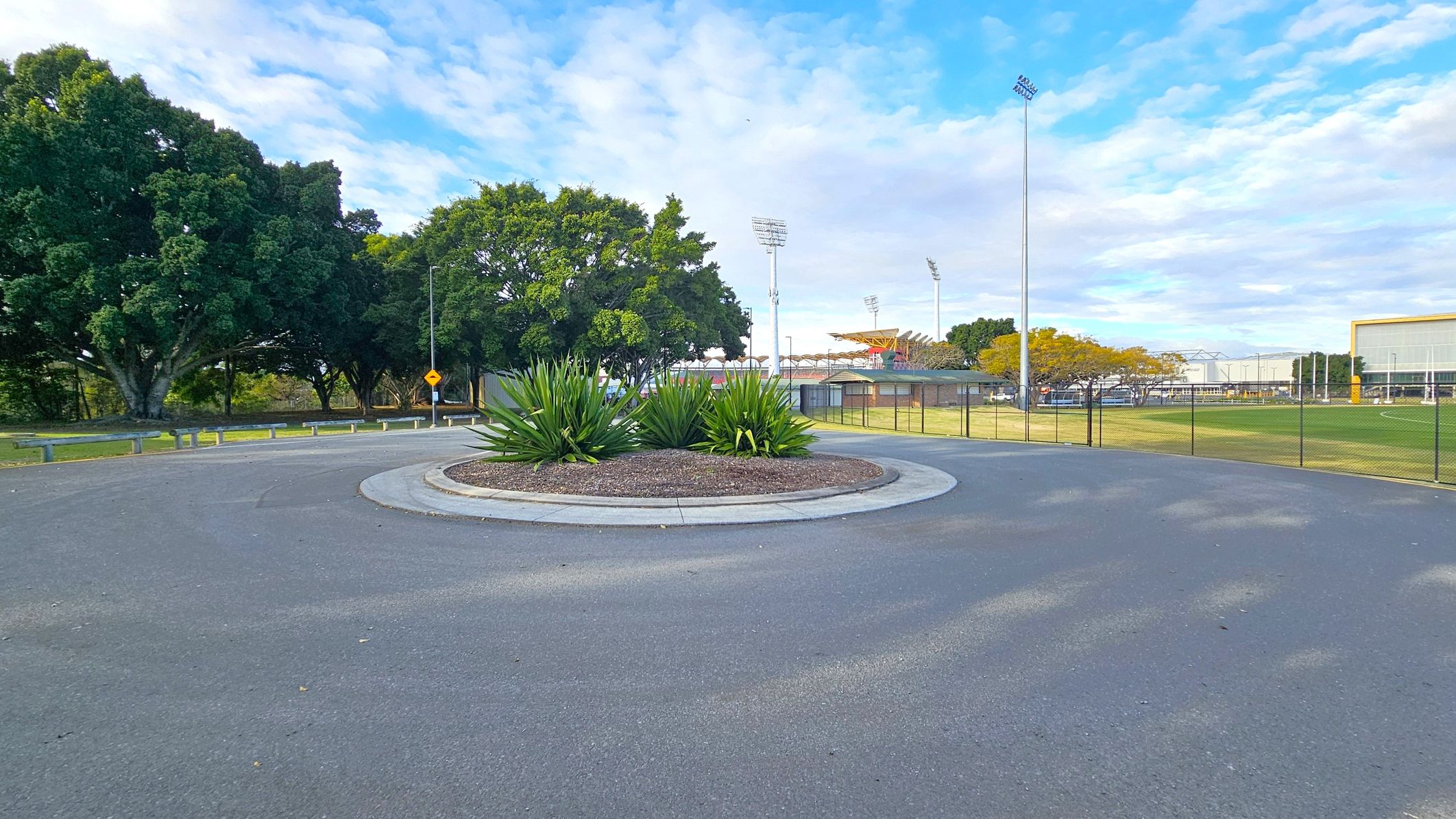 A roundabout with a central garden of green plants and mulch is in the foreground. To the left, there are large, leafy trees and a road with metal barriers. In the background, tall stadium lights and a sports field with a fence are visible. On the right, a building with a yellow trim and a grassy area can be seen. The sky is partly cloudy. A roundabout with a central garden of green plants and mulch is in the foreground. To the left, there are large, leafy trees and a road with metal barriers. In the background, tall stadium lights and a sports field with a fence are visible. On the right, a building with a yellow trim and a grassy area can be seen. The sky is partly cloudy.