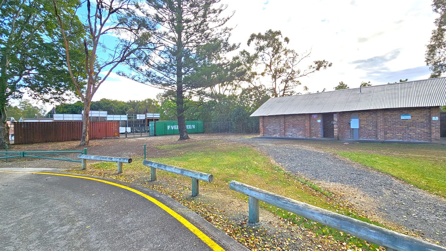 A paved road with a yellow line curves on the left, bordered by wooden barriers. To the right, a long brick building with a corrugated metal roof stands on a gravel path. In the centre, a large tree is surrounded by grass and scattered leaves. Behind the tree, shipping containers, including a green one marked "EVERGREEN," are visible. Tall trees form a backdrop, with a mix of green and brown foliage. A paved road with a yellow line curves on the left, bordered by wooden barriers. To the right, a long brick building with a corrugated metal roof stands on a gravel path. In the centre, a large tree is surrounded by grass and scattered leaves. Behind the tree, shipping containers, including a green one marked "EVERGREEN," are visible. Tall trees form a backdrop, with a mix of green and brown foliage.