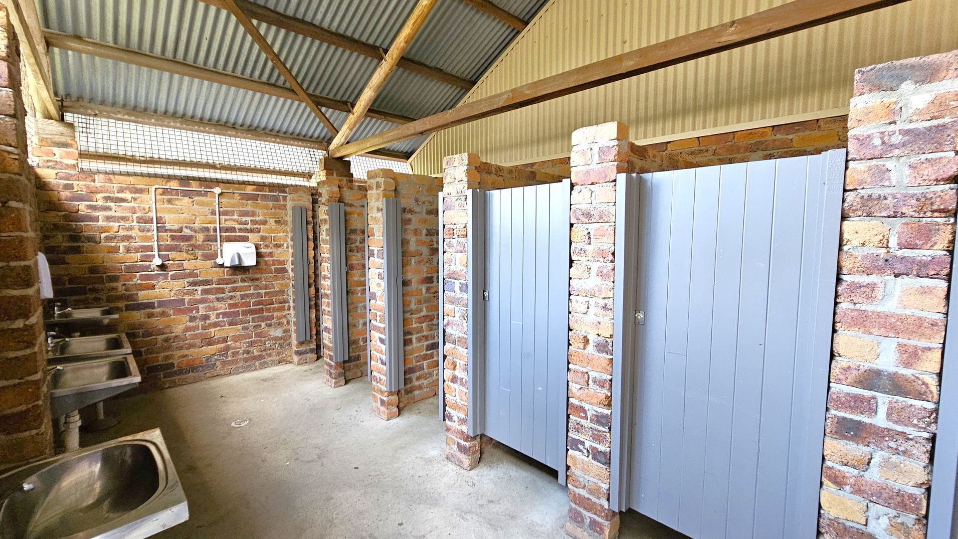 A public restroom with brick walls and a corrugated metal roof. On the left, there are several stainless steel sinks with a paper towel dispenser above. On the right, there are toilet cubicles with grey doors, separated by brick pillars. The floor is concrete, and the ceiling has exposed wooden beams. A public restroom with brick walls and a corrugated metal roof. On the left, there are several stainless steel sinks with a paper towel dispenser above. On the right, there are toilet cubicles with grey doors, separated by brick pillars. The floor is concrete, and the ceiling has exposed wooden beams.