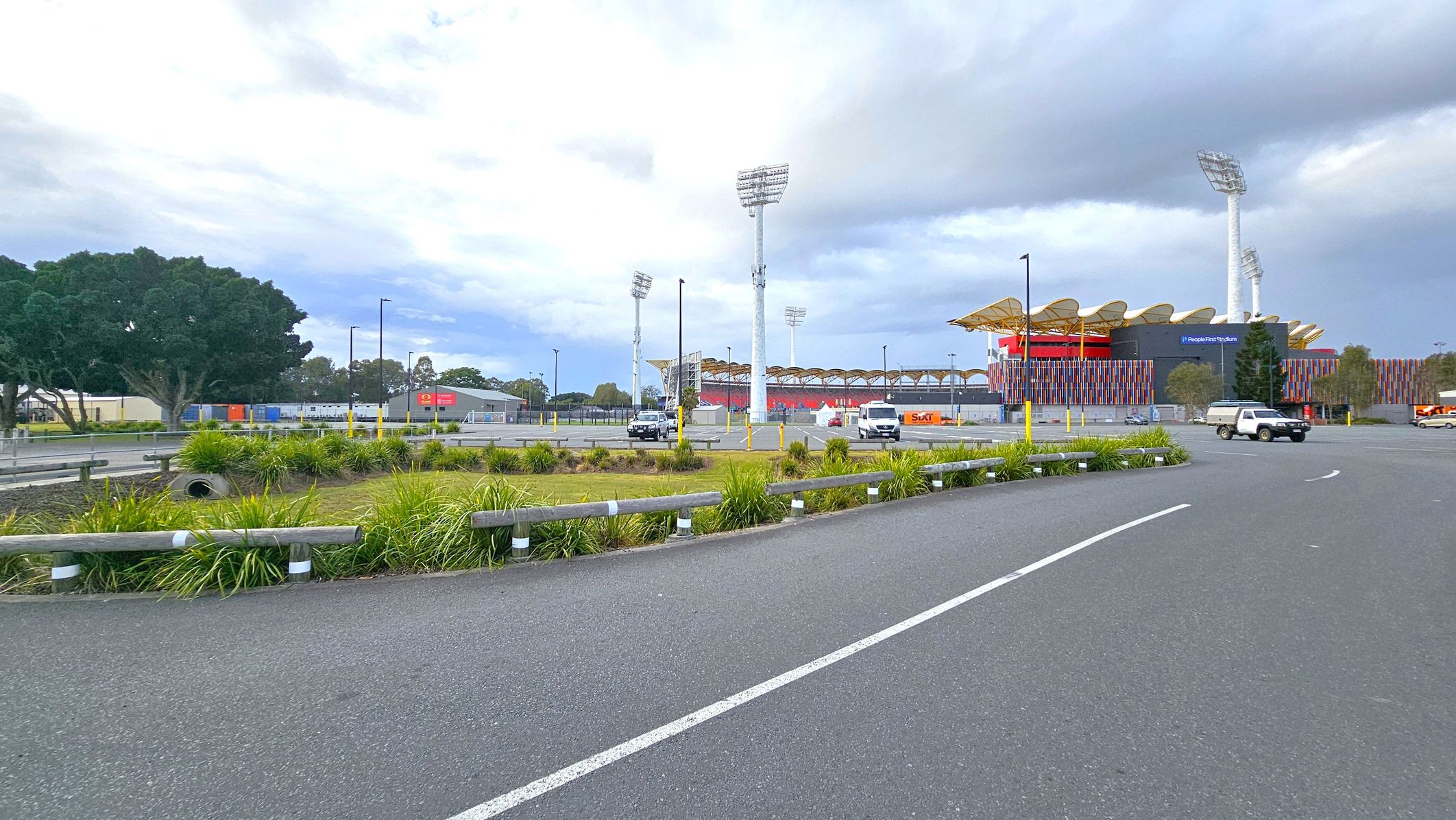 A large stadium with colourful vertical panels and a wavy roof is on the right, with tall floodlights surrounding it. In the foreground, there is a paved road with white lines and a grassy area with low bushes and a metal guardrail. Several vehicles, including a white van and a black car, are parked near the stadium. On the left, there are large trees and some industrial buildings with signage. The sky is overcast. A large stadium with colourful vertical panels and a wavy roof is on the right, with tall floodlights surrounding it. In the foreground, there is a paved road with white lines and a grassy area with low bushes and a metal guardrail. Several vehicles, including a white van and a black car, are parked near the stadium. On the left, there are large trees and some industrial buildings with signage. The sky is overcast.