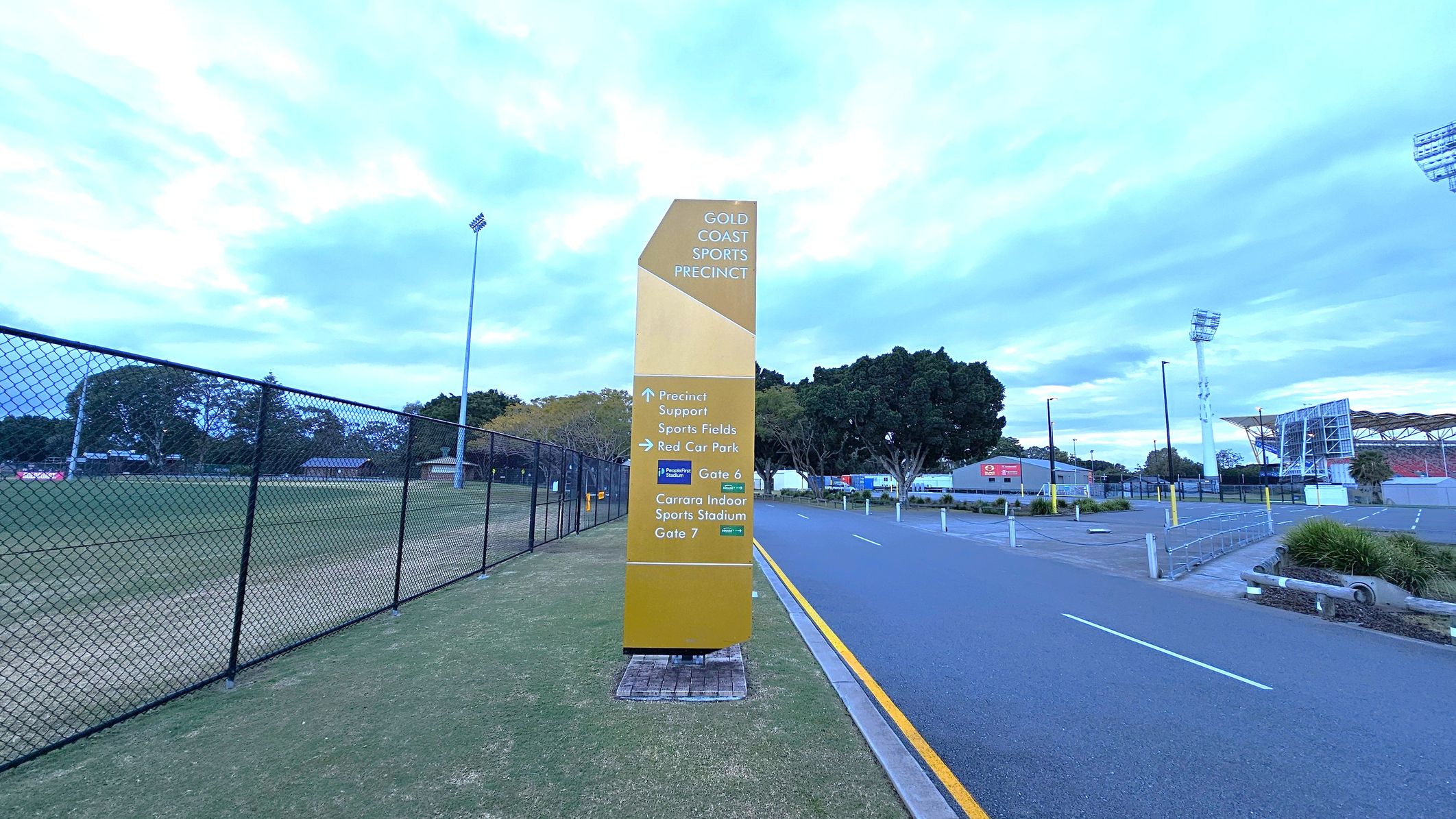 A tall, yellow sign stands in the centre, displaying directions for the Gold Coast Sports Precinct. On the left, a chain-link fence borders a grassy sports field with trees and a tall floodlight. On the right, a paved road with a yellow line runs alongside a car park and a stadium with a distinctive roof structure. Trees and a few buildings are visible in the background. A tall, yellow sign stands in the centre, displaying directions for the Gold Coast Sports Precinct. On the left, a chain-link fence borders a grassy sports field with trees and a tall floodlight. On the right, a paved road with a yellow line runs alongside a car park and a stadium with a distinctive roof structure. Trees and a few buildings are visible in the background.