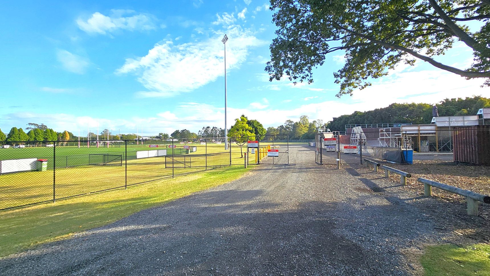 A gravel path leads to a fenced area with a gate in the centre. On the left, there's a green sports field with goalposts and a tall floodlight. A chain-link fence separates the field from the path. On the right, there are structures resembling bleachers or seating areas, with a blue bin nearby. Trees are visible in the background, and a large tree with overhanging branches is on the right. The sky is clear with some clouds. A gravel path leads to a fenced area with a gate in the centre. On the left, there's a green sports field with goalposts and a tall floodlight. A chain-link fence separates the field from the path. On the right, there are structures resembling bleachers or seating areas, with a blue bin nearby. Trees are visible in the background, and a large tree with overhanging branches is on the right. The sky is clear with some clouds.