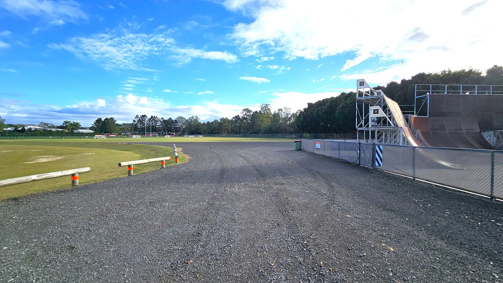 A gravel path runs through the centre, bordered by a grassy field on the left with wooden barriers and orange cones. On the right, a large skate ramp structure with metal railings and platforms is visible, enclosed by a chain-link fence. In the background, there are trees and a few buildings, with a clear blue sky above. A gravel path runs through the centre, bordered by a grassy field on the left with wooden barriers and orange cones. On the right, a large skate ramp structure with metal railings and platforms is visible, enclosed by a chain-link fence. In the background, there are trees and a few buildings, with a clear blue sky above.