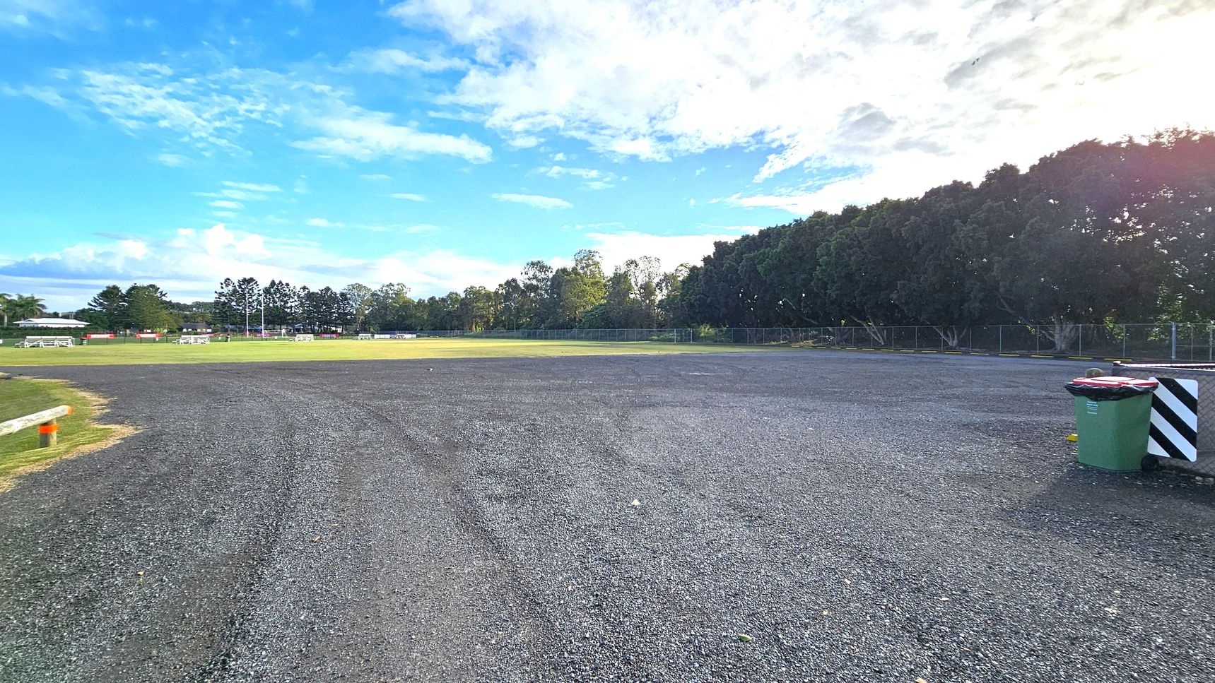 A gravel area in the foreground with a green rubbish bin on the right. In the background, there's a grassy field with white goalposts and benches. Trees line the far edge of the field, and a fence runs along the perimeter. The sky is partly cloudy. A gravel area in the foreground with a green rubbish bin on the right. In the background, there's a grassy field with white goalposts and benches. Trees line the far edge of the field, and a fence runs along the perimeter. The sky is partly cloudy.