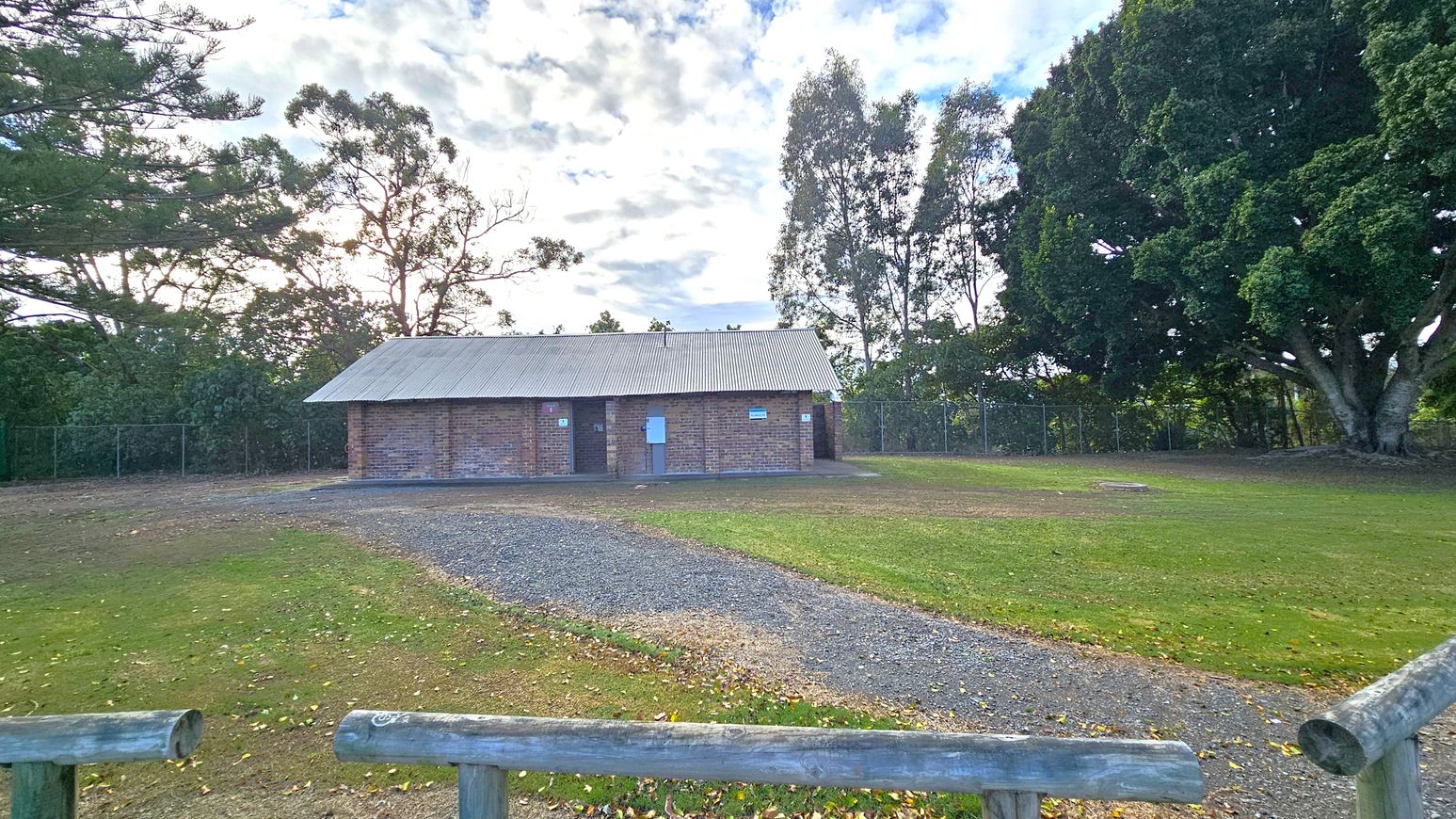 A small brick building with a corrugated metal roof is in the centre, surrounded by a grassy area. A gravel path leads up to the building. On the right, there is a large tree with dense foliage. On the left, there are more trees and a wooden fence in the foreground. The background is filled with more trees and a partly cloudy sky. A small brick building with a corrugated metal roof is in the centre, surrounded by a grassy area. A gravel path leads up to the building. On the right, there is a large tree with dense foliage. On the left, there are more trees and a wooden fence in the foreground. The background is filled with more trees and a partly cloudy sky.