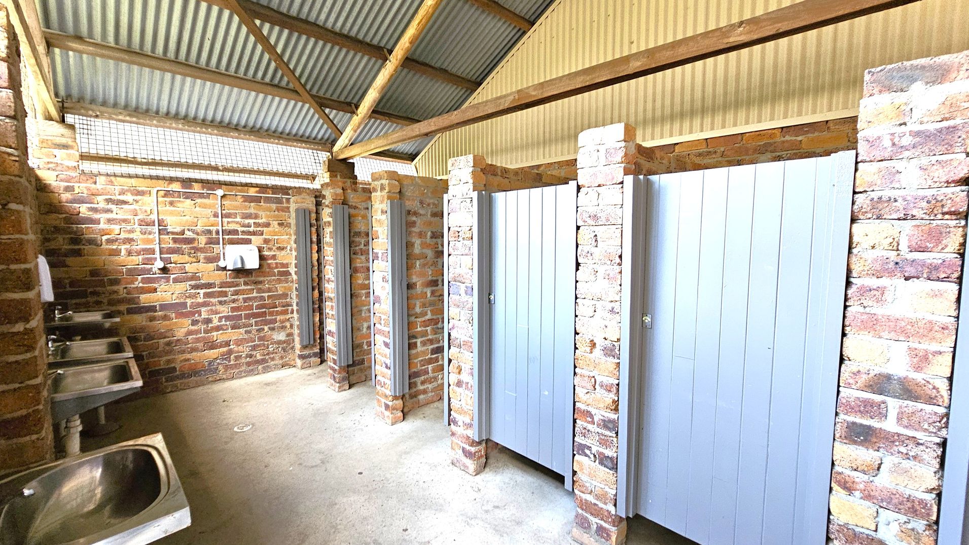 A public restroom with brick walls and a corrugated metal roof. On the left, there are three stainless steel sinks with a soap dispenser and a hand dryer mounted on the wall. On the right, there are several toilet cubicles with light grey doors, separated by brick pillars. The floor is concrete, and the ceiling has exposed wooden beams. A public restroom with brick walls and a corrugated metal roof. On the left, there are three stainless steel sinks with a soap dispenser and a hand dryer mounted on the wall. On the right, there are several toilet cubicles with light grey doors, separated by brick pillars. The floor is concrete, and the ceiling has exposed wooden beams.