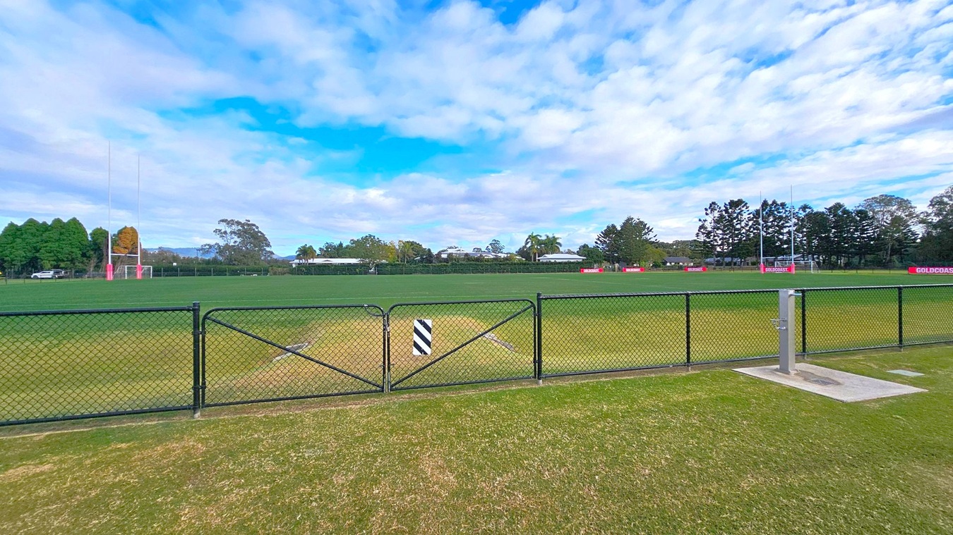 A grassy sports field with goalposts on both the left and right sides. A black chain-link fence runs across the foreground, with a small gate in the centre. Behind the field, there are trees and a few low buildings. The grass is well-maintained, and there are signs with "GOLDCOAST" written on them along the perimeter. The sky is partly cloudy. A grassy sports field with goalposts on both the left and right sides. A black chain-link fence runs across the foreground, with a small gate in the centre. Behind the field, there are trees and a few low buildings. The grass is well-maintained, and there are signs with "GOLDCOAST" written on them along the perimeter. The sky is partly cloudy.