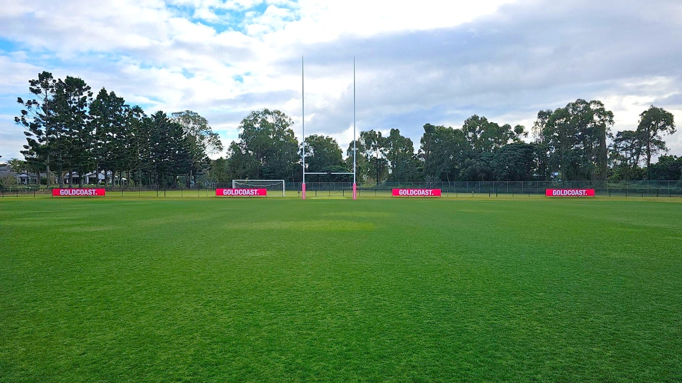 A rugby field with lush green grass. In the centre, there are rugby goalposts with a football goal behind them. Red advertising boards with "GOLDCOAST." are placed along the fence. Tall trees form a dense line in the background, providing a natural border. The sky is partly cloudy, casting a soft light over the scene. A rugby field with lush green grass. In the centre, there are rugby goalposts with a football goal behind them. Red advertising boards with "GOLDCOAST." are placed along the fence. Tall trees form a dense line in the background, providing a natural border. The sky is partly cloudy, casting a soft light over the scene.