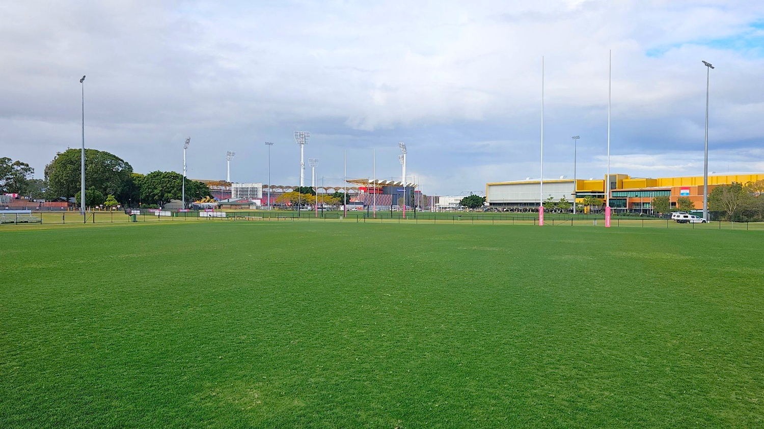 A large, open grassy field with rugby posts in the centre. In the background, there are several tall floodlights and a mix of trees and buildings. On the right, a large yellow and grey building is visible, with a white vehicle parked nearby. The field is surrounded by a low fence, and there are benches and a bin on the left side. A large, open grassy field with rugby posts in the centre. In the background, there are several tall floodlights and a mix of trees and buildings. On the right, a large yellow and grey building is visible, with a white vehicle parked nearby. The field is surrounded by a low fence, and there are benches and a bin on the left side.