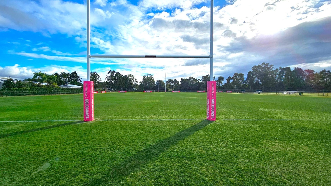A rugby field with goalposts in the foreground, featuring padding labelled "Australia's Gold Coast" on each post. The grass is well-maintained, and the field extends into the distance. In the background, there are trees and some low buildings, with a partly cloudy sky above. Shadows from the goalposts stretch across the grass. A rugby field with goalposts in the foreground, featuring padding labelled "Australia's Gold Coast" on each post. The grass is well-maintained, and the field extends into the distance. In the background, there are trees and some low buildings, with a partly cloudy sky above. Shadows from the goalposts stretch across the grass.
