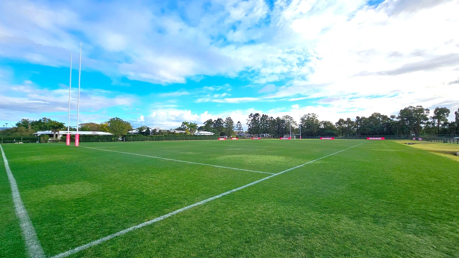A rugby field with lush green grass and white boundary lines. On the left, there are tall goalposts with pink padding at the base. In the background, there are trees and a few low buildings. On the right, another set of goalposts is visible, and there are red advertising boards along the perimeter. The field is expansive and well-maintained. A rugby field with lush green grass and white boundary lines. On the left, there are tall goalposts with pink padding at the base. In the background, there are trees and a few low buildings. On the right, another set of goalposts is visible, and there are red advertising boards along the perimeter. The field is expansive and well-maintained.