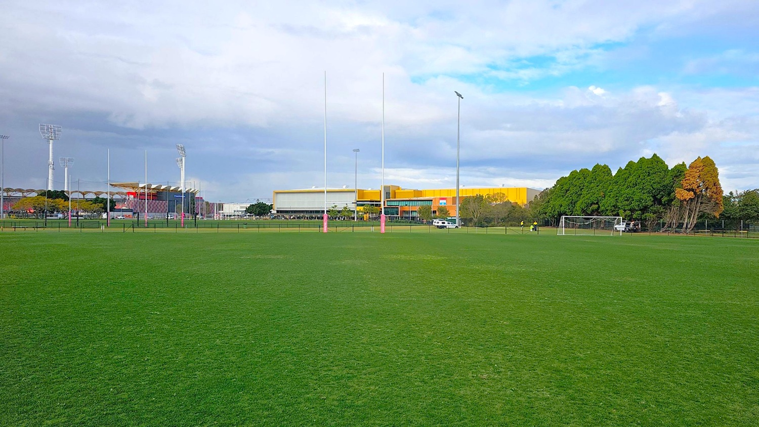 A sports field with green grass in the foreground. In the middle, there are rugby posts with pink padding. To the right, a football goal is visible near a line of trees with some autumn foliage. In the background, there are large buildings with a mix of yellow and grey colours, and several tall floodlights. A white vehicle is parked near the buildings. A sports field with green grass in the foreground. In the middle, there are rugby posts with pink padding. To the right, a football goal is visible near a line of trees with some autumn foliage. In the background, there are large buildings with a mix of yellow and grey colours, and several tall floodlights. A white vehicle is parked near the buildings.