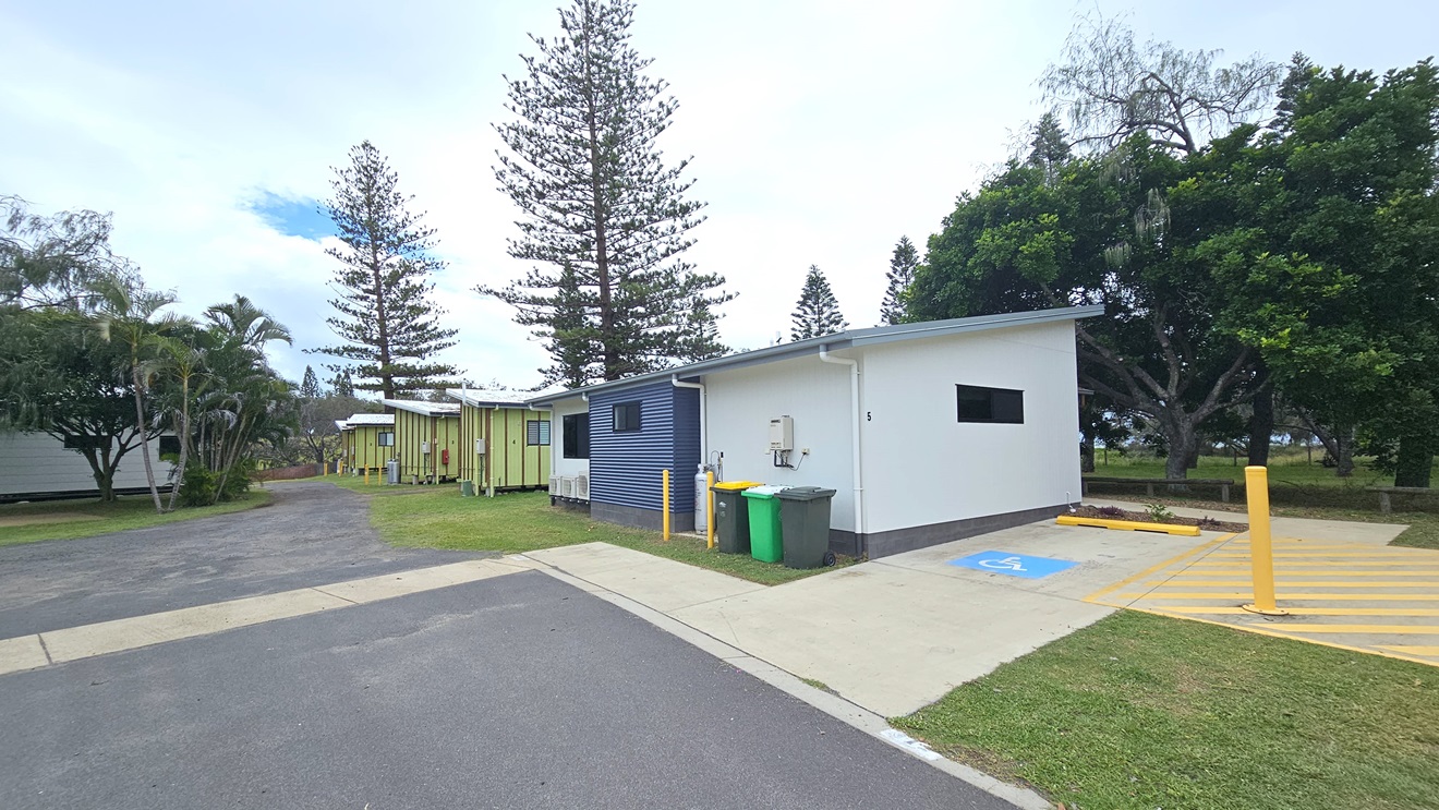 The accessible cabin is nestled on green grass near the park. Accessible parking, marked by a blue wheelchair sign and yellow diagonal lines with a bollard, is on the right. Three rubbish bins are at the back. To the left, the road leads to other cabins, while another facility building is nearby. On the right is a park with pine and palm trees. The accessible cabin is nestled on green grass near the park. Accessible parking, marked by a blue wheelchair sign and yellow diagonal lines with a bollard, is on the right. Three rubbish bins are at the back. To the left, the road leads to other cabins, while another facility building is nearby. On the right is a park with pine and palm trees.