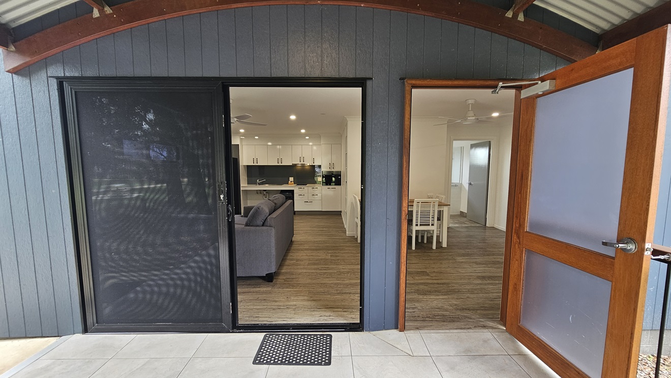 There are two entrance doors. The right one is a manual sliding glass door, equipped with a non-slippery mat while the left door is a manual outward opening door. Inside, the floor is timber, and the walls and ceiling are white with ceiling fans installed. The kitchen area, dining table, and a grey couch are also visible. Outside, the entrance features an arched ceiling with a corrugated roof, and the wall has grey wooden planks. The floor has grey tiles. There are two entrance doors. The right one is a manual sliding glass door, equipped with a non-slippery mat while the left door is a manual outward opening door. Inside, the floor is timber, and the walls and ceiling are white with ceiling fans installed. The kitchen area, dining table, and a grey couch are also visible. Outside, the entrance features an arched ceiling with a corrugated roof, and the wall has grey wooden planks. The floor has grey tiles.