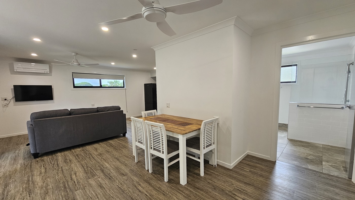 Inside the cabin, timber floors complement white walls and ceilings adorned with a ceiling fan and LED lights. A grey couch sits to the left, alongside a wooden dining set. A flatscreen TV and air conditioning are mounted on the left wall. Next to them, a glass window offers natural light, with a fridge nearby. To the right, a grey door leads to the bathroom, showcasing a shower with handgrabs, white walls, and grey tiles. Inside the cabin, timber floors complement white walls and ceilings adorned with a ceiling fan and LED lights. A grey couch sits to the left, alongside a wooden dining set. A flatscreen TV and air conditioning are mounted on the left wall. Next to them, a glass window offers natural light, with a fridge nearby. To the right, a grey door leads to the bathroom, showcasing a shower with handgrabs, white walls, and grey tiles.