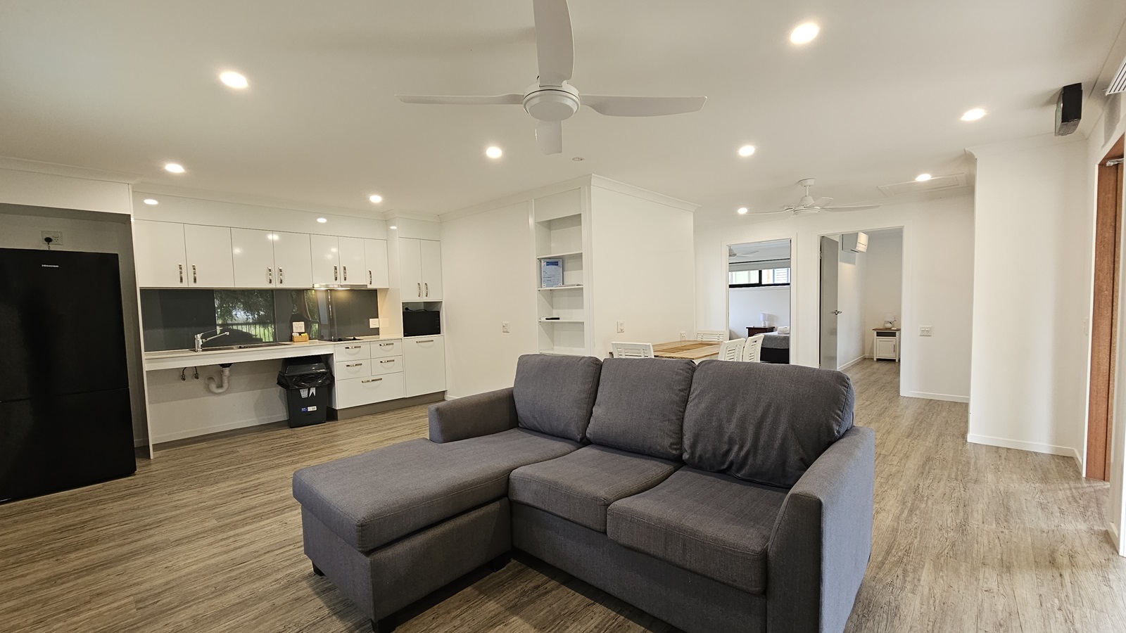 In the centre is an L-shaped grey couch. To the left is the kitchen area, equipped with cabinets, sink, drawers, microwave oven, and a fridge. Adjacent to the couch is a dining table. To the right, a door leads to the bedroom. The floor is timber, with white walls and ceiling featuring LED lights and ceiling fans. In the centre is an L-shaped grey couch. To the left is the kitchen area, equipped with cabinets, sink, drawers, microwave oven, and a fridge. Adjacent to the couch is a dining table. To the right, a door leads to the bedroom. The floor is timber, with white walls and ceiling featuring LED lights and ceiling fans.