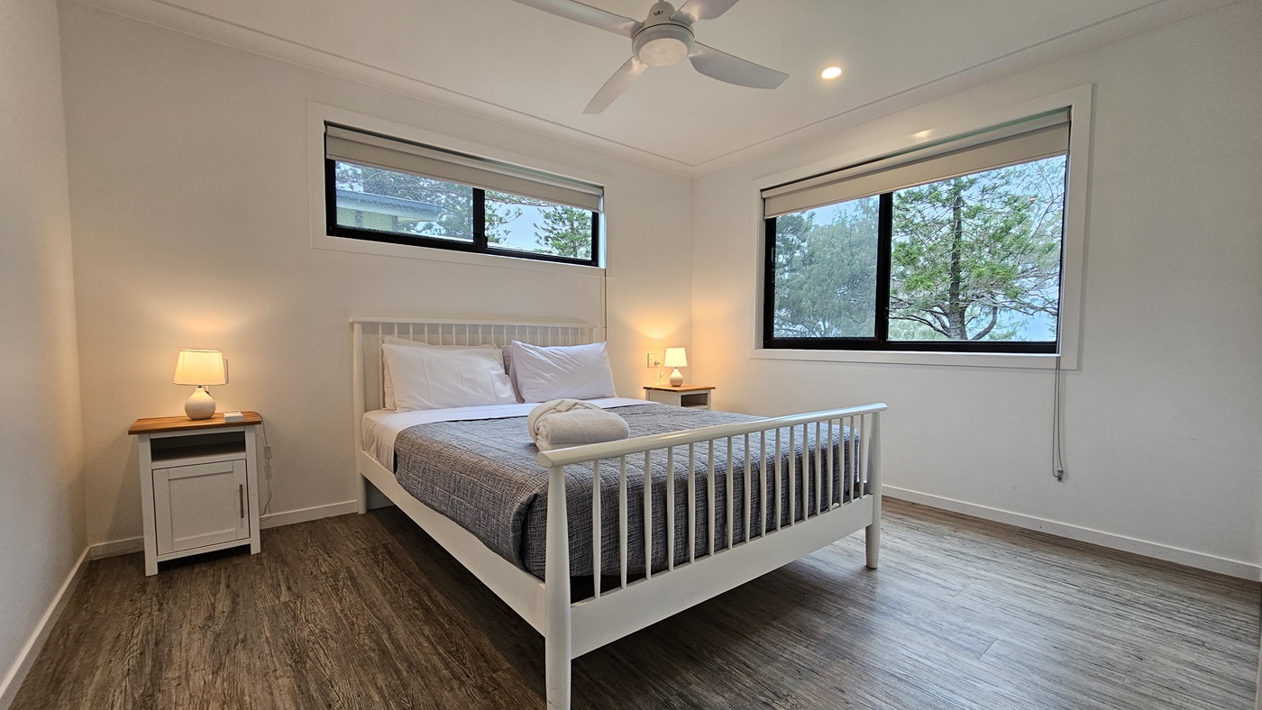 In the centre stands a white queen-size bed adorned with a grey cover. White pillows and a neatly rolled white towel rest atop. Flanking the bed are two bedside tables, each with a lampshade. A window is situated on the central and right walls. The walls and ceiling are painted white, illuminated by LED lights and a ceiling fan. The floor is timber. In the centre stands a white queen-size bed adorned with a grey cover. White pillows and a neatly rolled white towel rest atop. Flanking the bed are two bedside tables, each with a lampshade. A window is situated on the central and right walls. The walls and ceiling are painted white, illuminated by LED lights and a ceiling fan. The floor is timber.