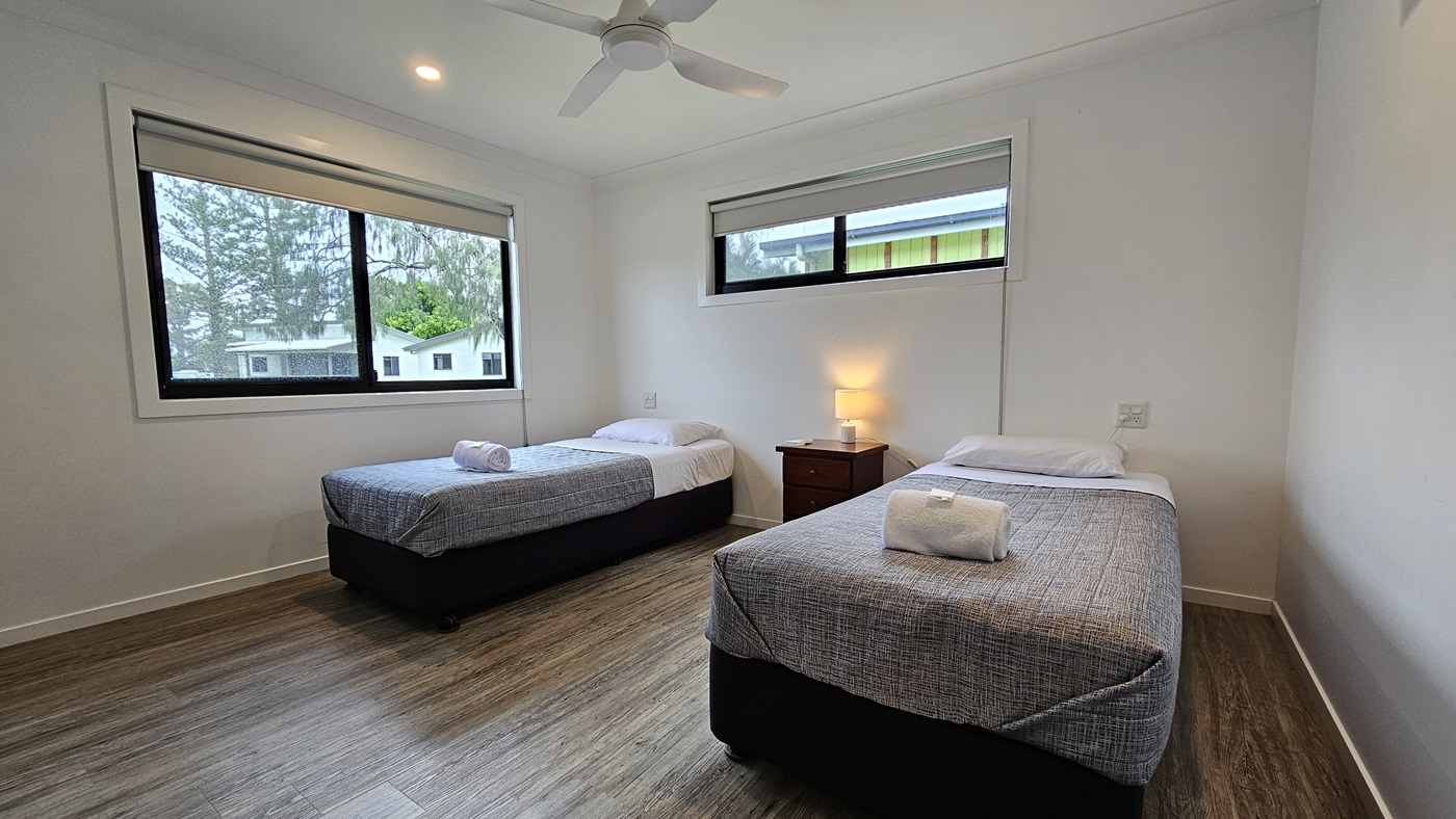 In the room, two single beds with grey covers stand opposite each other. White pillows and a neatly rolled white towel sit atop each bed. A window is positioned on the middle and right walls. Between the beds, a bedside table hosts a lampshade. The walls and ceiling are painted white, illuminated by LED lights and a ceiling fan. The floor is timber. Outside the window, trees and other cabins are visible. In the room, two single beds with grey covers stand opposite each other. White pillows and a neatly rolled white towel sit atop each bed. A window is positioned on the middle and right walls. Between the beds, a bedside table hosts a lampshade. The walls and ceiling are painted white, illuminated by LED lights and a ceiling fan. The floor is timber. Outside the window, trees and other cabins are visible.