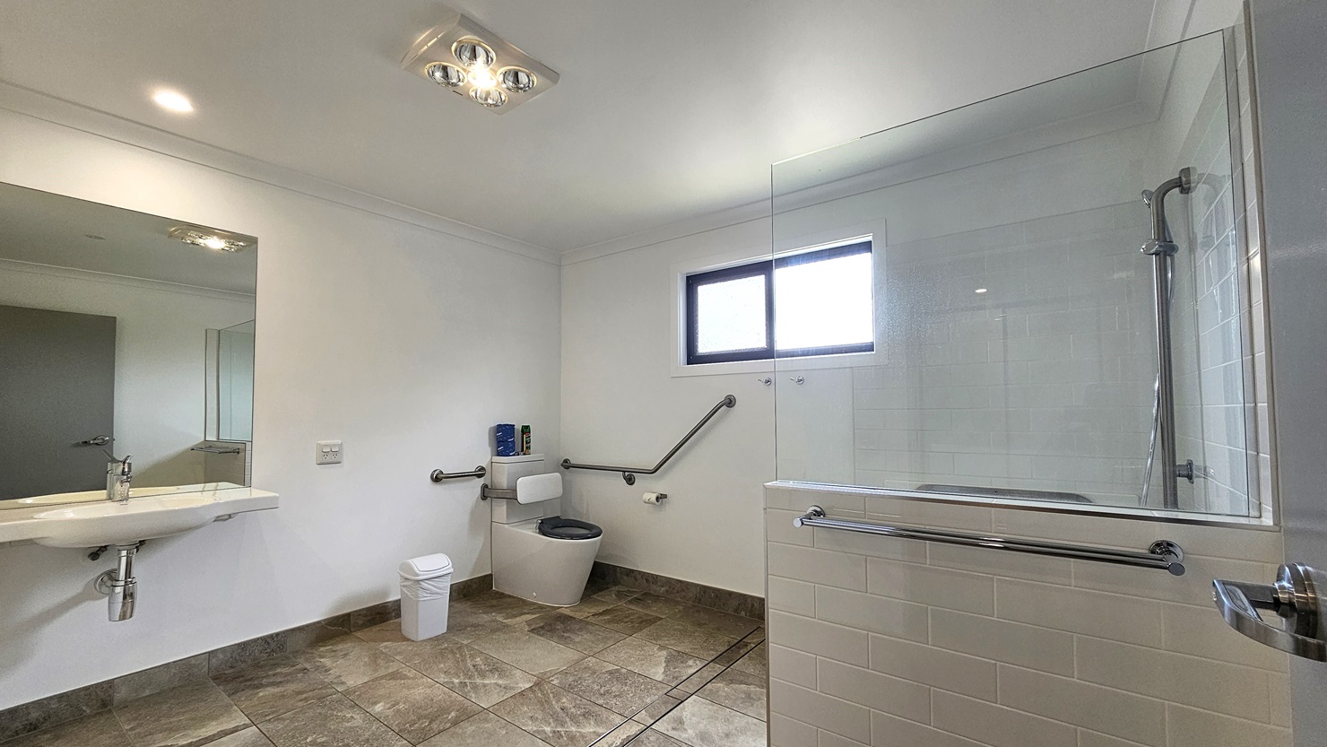 In the accessible bathroom, a toilet bowl with hand grabs is situated in the left corner, accompanied by a rubbish bin and a tissue dispenser. Adjacent to this is a hand basin with a mirror mounted on the glass wall. To the right, a shower is present, separated by a combination of glass and grey brick tiles. The floor features grey tiles with a marble design. The walls are white, and the ceiling is white with LED lights. In the accessible bathroom, a toilet bowl with hand grabs is situated in the left corner, accompanied by a rubbish bin and a tissue dispenser. Adjacent to this is a hand basin with a mirror mounted on the glass wall. To the right, a shower is present, separated by a combination of glass and grey brick tiles. The floor features grey tiles with a marble design. The walls are white, and the ceiling is white with LED lights.