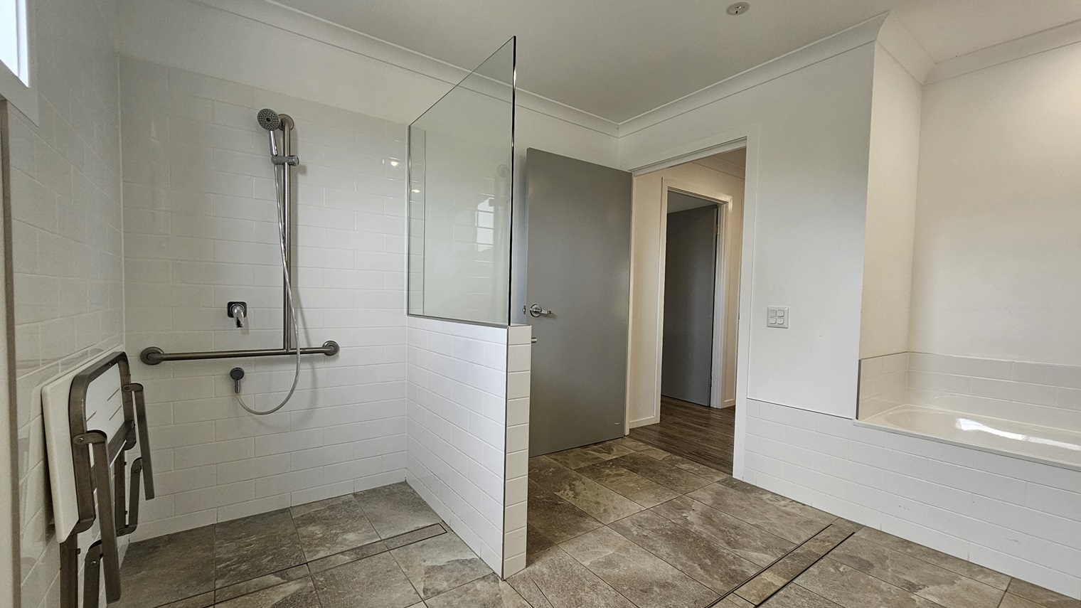 In the accessible bathroom, a shower with a handrail and foldable bench is on the left, separated by a partition of glass and grey brick tiles. On the right is a bath tub against the wall. The floor boasts grey tiles with a marble design. The walls and the ceiling are white with LED lights. In the accessible bathroom, a shower with a handrail and foldable bench is on the left, separated by a partition of glass and grey brick tiles. On the right is a bath tub against the wall. The floor boasts grey tiles with a marble design. The walls and the ceiling are white with LED lights.