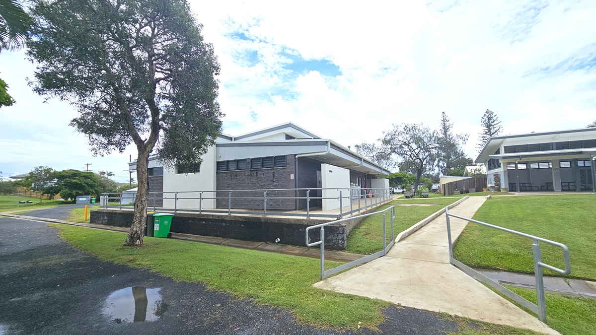 In a grassy area stands a low-rise facility with a grey brick facade. A brick pathway leads to it, alongside a step-free ramp with railings. Four gas tanks are displayed in front of the building. To the left is a round circular structure, and to the right is a wooden planter box with plants. Trees surround the area. In a grassy area stands a low-rise facility with a grey brick facade. A brick pathway leads to it, alongside a step-free ramp with railings. Four gas tanks are displayed in front of the building. To the left is a round circular structure, and to the right is a wooden planter box with plants. Trees surround the area.