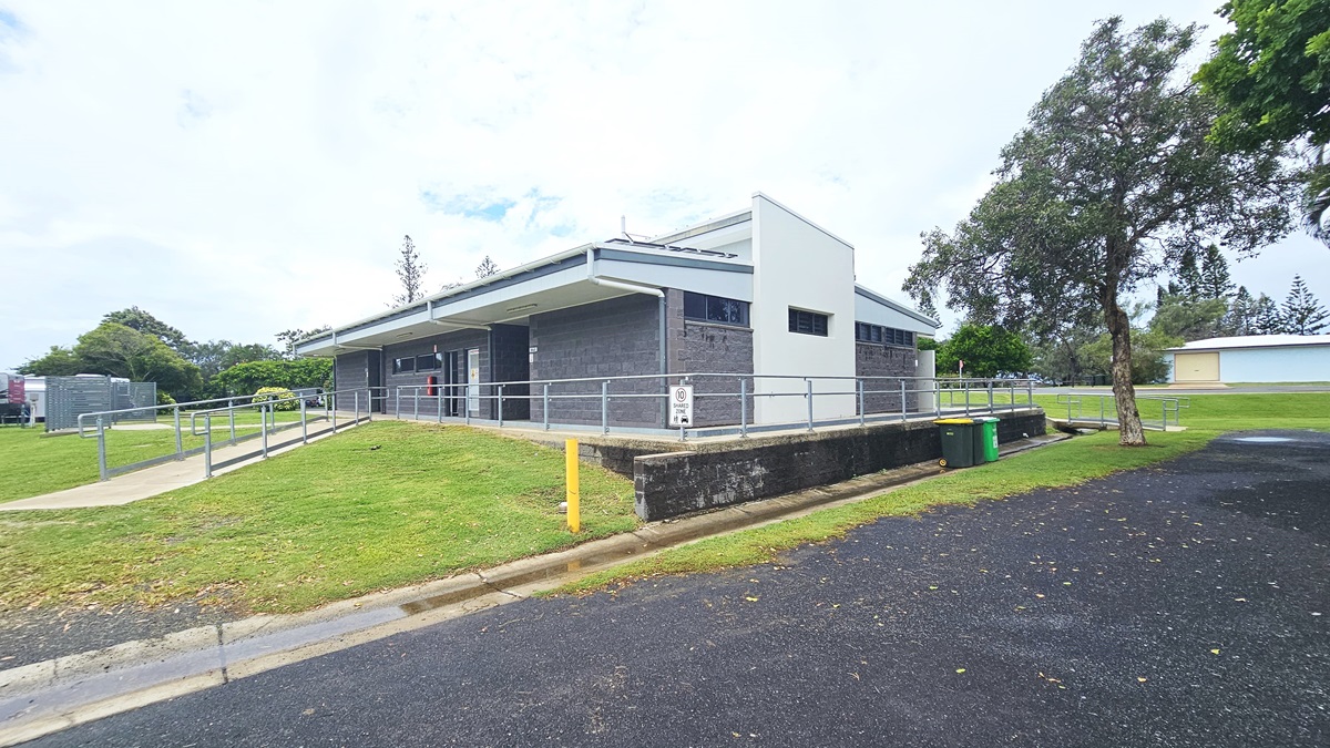 In the middle stands a low-rise facility with a dark grey brick facade. An accessible ramp entry with handrails is on the left, with an asphalt road to the right. Two rubbish bins are placed near the concrete wall. Surrounding the area are trees, grass, and other visible building structures in the distance. In the middle stands a low-rise facility with a dark grey brick facade. An accessible ramp entry with handrails is on the left, with an asphalt road to the right. Two rubbish bins are placed near the concrete wall. Surrounding the area are trees, grass, and other visible building structures in the distance.