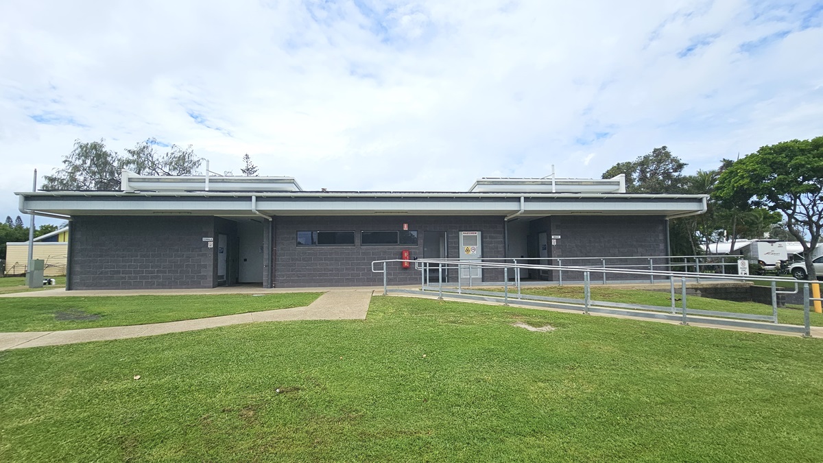 In the middle stands a low-rise facility with a dark grey brick facade, featuring small windows and multiple doors. An accessible ramp entry with handrails is on the right. To the left, brick pathways cut through the grass and lead to the building. Surrounding the area are trees, grass, and other visible building structures in the distance. In the middle stands a low-rise facility with a dark grey brick facade, featuring small windows and multiple doors. An accessible ramp entry with handrails is on the right. To the left, brick pathways cut through the grass and lead to the building. Surrounding the area are trees, grass, and other visible building structures in the distance.