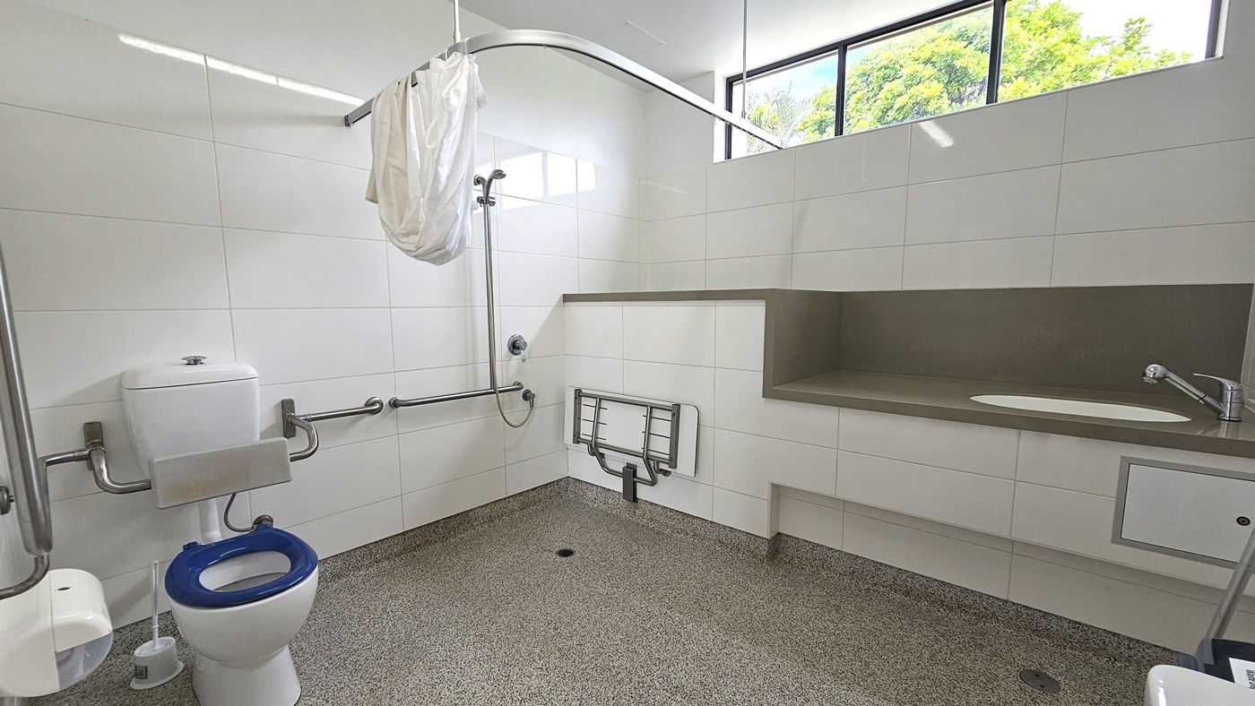 Inside the accessible bathroom, a toilet bowl with hand grabs, a tissue dispenser, and a toilet brush are on the left. On the right, a shower with handrails and curtain, alongside a foldable bench chair mounted to the wall. Adjacent, is a lavatory with a sink. The walls feature white tiles with glass windows. Floor tiles have a granite design. Inside the accessible bathroom, a toilet bowl with hand grabs, a tissue dispenser, and a toilet brush are on the left. On the right, a shower with handrails and curtain, alongside a foldable bench chair mounted to the wall. Adjacent, is a lavatory with a sink. The walls feature white tiles with glass windows. Floor tiles have a granite design.
