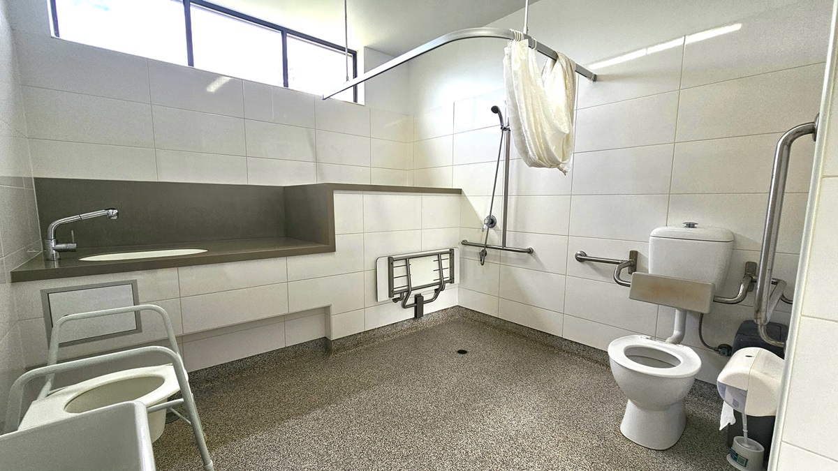 Inside the accessible bathroom, a toilet bowl with hand grabs, a tissue dispenser, a rubbish bin, and a toilet brush are on the right. On the left is a shower with handrails and curtain and a foldable bench chair mounted to the wall. Adjacent, is a lavatory with a sink. The walls feature white tiles with glass windows. A toilet pot is positioned on the left, near a rubbish bin. Floor tiles have a granite design. Inside the accessible bathroom, a toilet bowl with hand grabs, a tissue dispenser, a rubbish bin, and a toilet brush are on the right. On the left is a shower with handrails and curtain and a foldable bench chair mounted to the wall. Adjacent, is a lavatory with a sink. The walls feature white tiles with glass windows. A toilet pot is positioned on the left, near a rubbish bin. Floor tiles have a granite design.