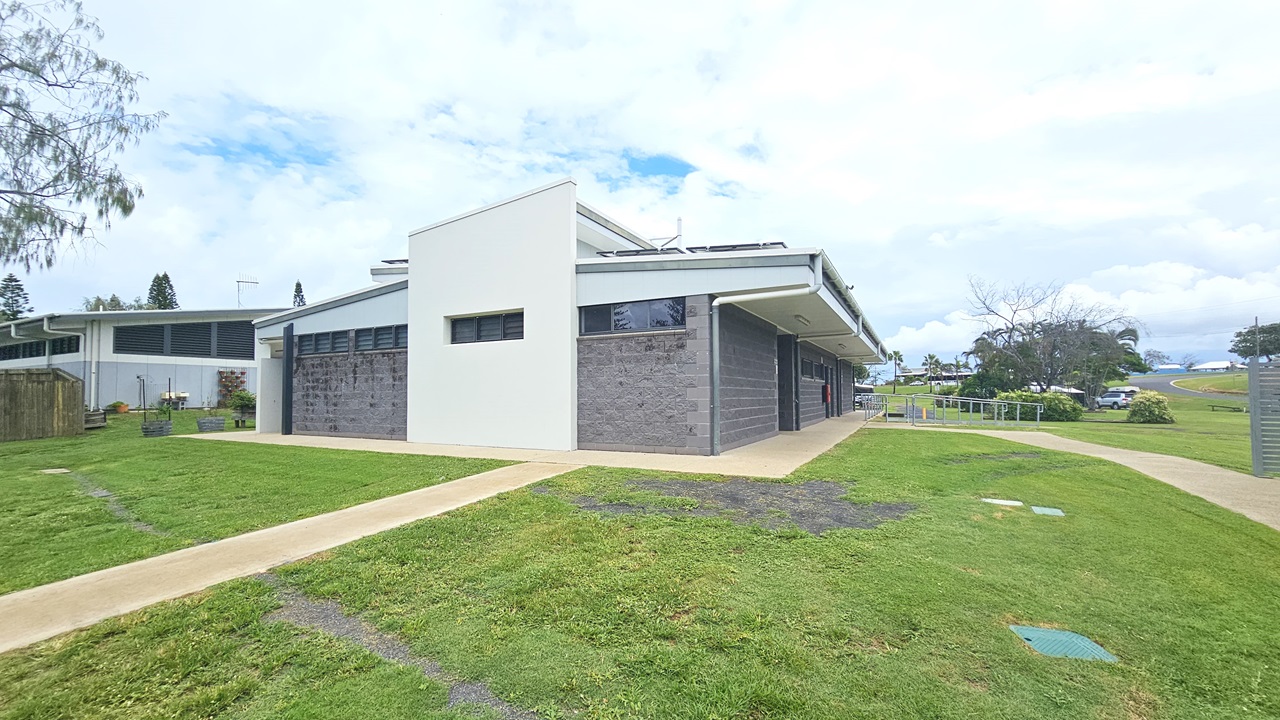 In the middle stands a low-rise facility with a dark grey brick facade, featuring small windows. An accessible ramp entry with handrails is on the right. Brick pathways cut through the grass, leading to the building. To the left is another building structure and planter boxes. Surrounding the area are trees and grass. Vehicles are parked under the shade of trees visible in the distance. In the middle stands a low-rise facility with a dark grey brick facade, featuring small windows. An accessible ramp entry with handrails is on the right. Brick pathways cut through the grass, leading to the building. To the left is another building structure and planter boxes. Surrounding the area are trees and grass. Vehicles are parked under the shade of trees visible in the distance.