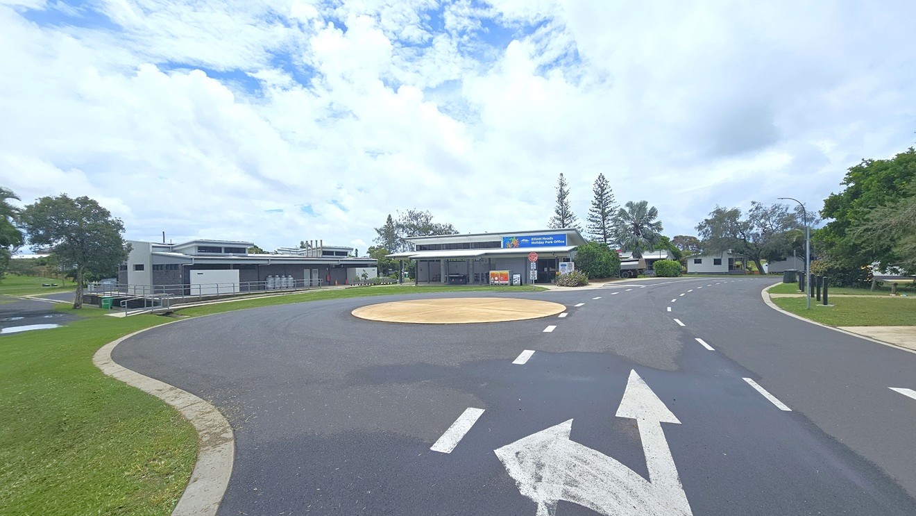 A single-storey building with a flat roof and a sign reading "Elliott Heads Holiday Park Office" is in the centre. The building has an open area with tables and chairs. In front, there are signs including a red "Guests Only Past This Point" and a "10 Shared Zone" sign. The foreground features a grassy area and a paved path leading to the entrance. Trees and additional buildings are visible in the background. A single-storey building with a flat roof and a sign reading "Elliott Heads Holiday Park Office" is in the centre. The building has an open area with tables and chairs. In front, there are signs including a red "Guests Only Past This Point" and a "10 Shared Zone" sign. The foreground features a grassy area and a paved path leading to the entrance. Trees and additional buildings are visible in the background.