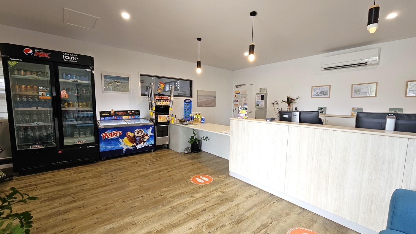 A reception desk with a timber front holds computers on the right. Adjacent is a lower desk with brochures. To the left is a fridge displaying cold drinks and an ice cream freezer, along with a drinks dispenser. The white ceiling and wall feature light fixtures, posters, and decorations. The timber-tiled floor is decorated with a potted plant on the left, and a blue couch is positioned in front of the reception desk. A reception desk with a timber front holds computers on the right. Adjacent is a lower desk with brochures. To the left is a fridge displaying cold drinks and an ice cream freezer, along with a drinks dispenser. The white ceiling and wall feature light fixtures, posters, and decorations. The timber-tiled floor is decorated with a potted plant on the left, and a blue couch is positioned in front of the reception desk.