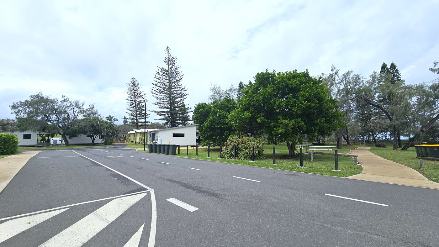 The road features white road markings, bordered by black bollards. A concrete pathway leads to the park on the right, amidst trees and grass. Rubbish bins line the road's edge. In the distance, cabins and a tall street light are visible. The road features white road markings, bordered by black bollards. A concrete pathway leads to the park on the right, amidst trees and grass. Rubbish bins line the road's edge. In the distance, cabins and a tall street light are visible.