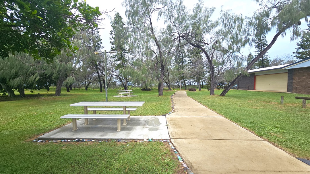 A concrete pathway cuts through grass, leading to picnic tables on the left, set on a concrete floor amidst grass. On the right is a low-rise building with a brown brick facade. Various trees and grass surround the area. A concrete pathway cuts through grass, leading to picnic tables on the left, set on a concrete floor amidst grass. On the right is a low-rise building with a brown brick facade. Various trees and grass surround the area.