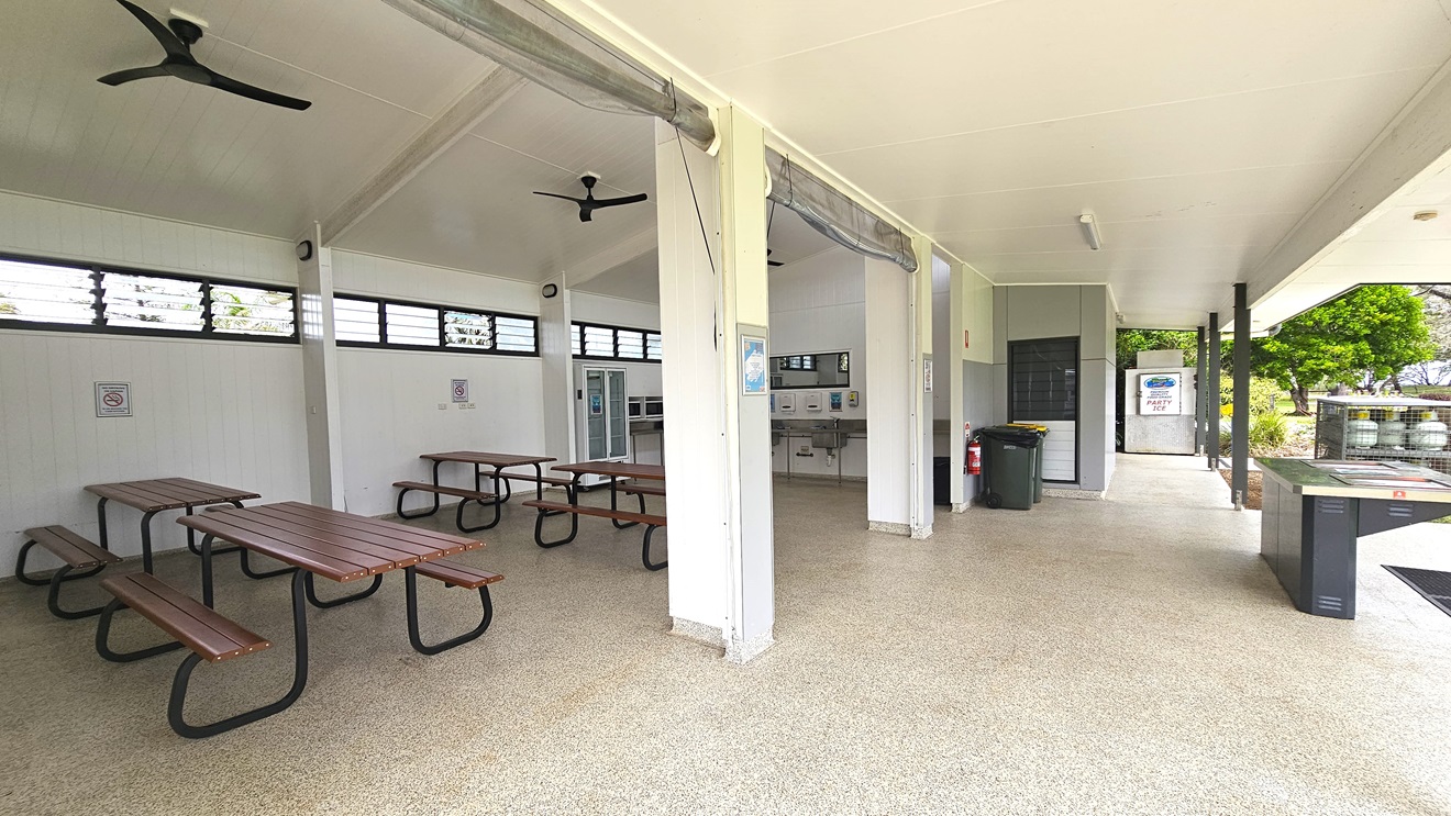 The BBQ area is spacious with picnic tables on the sides and a fridge. A sink and mirror are in the middle wall, and to the right is a barbecue grill. Gas tanks are in a cage nearby, with two rubbish bins and a fire extinguisher on the outer wall. White ceilings and walls feature ceiling fans and jalousy windows for ventilation. Safety reminders are affixed to the walls. Trees are visible on the right. The BBQ area is spacious with picnic tables on the sides and a fridge. A sink and mirror are in the middle wall, and to the right is a barbecue grill. Gas tanks are in a cage nearby, with two rubbish bins and a fire extinguisher on the outer wall. White ceilings and walls feature ceiling fans and jalousy windows for ventilation. Safety reminders are affixed to the walls. Trees are visible on the right.