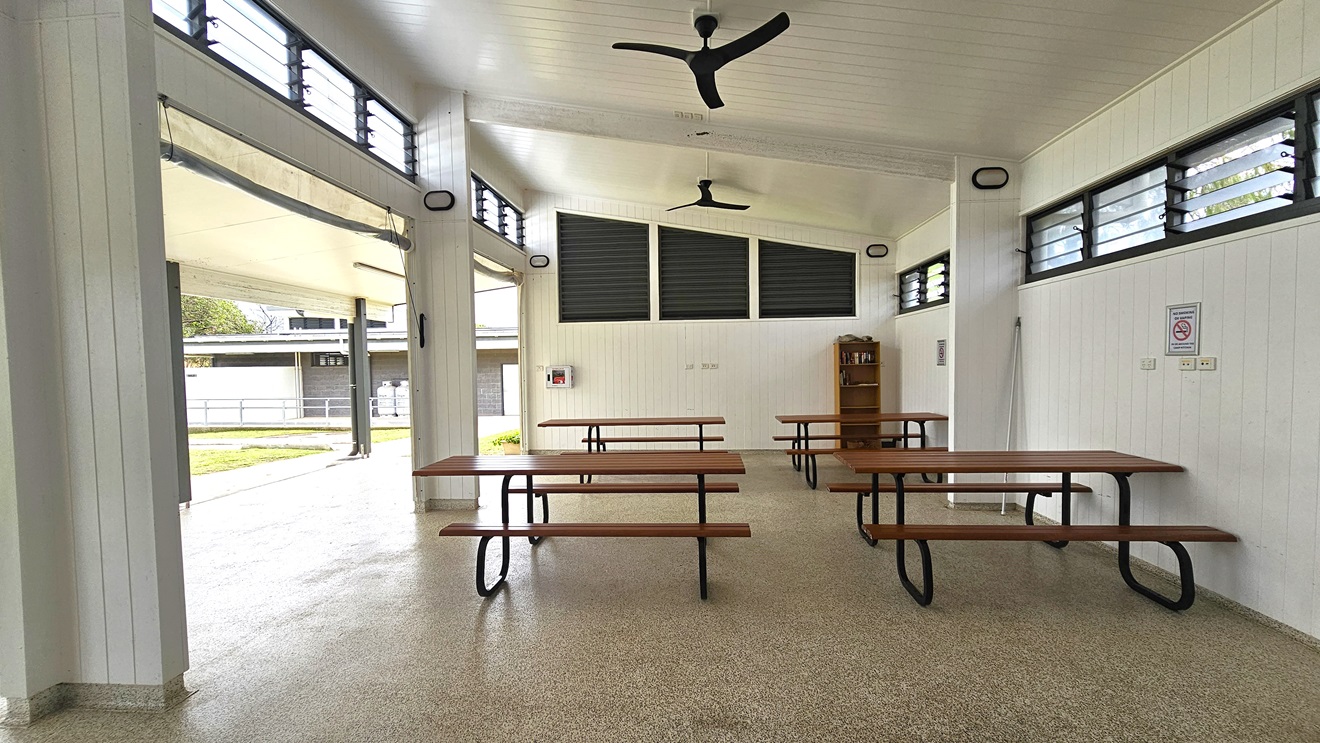 The BBQ area is spacious with picnic tables and a wooden display rack in the right corner. The pitched white ceiling has ceiling fans. The walls feature a white timber design, with jalousy windows and light fixtures on the upper wall. Safety reminders are affixed to walls. To the left is a low-rise facility building nestled on the green grass. The BBQ area is spacious with picnic tables and a wooden display rack in the right corner. The pitched white ceiling has ceiling fans. The walls feature a white timber design, with jalousy windows and light fixtures on the upper wall. Safety reminders are affixed to walls. To the left is a low-rise facility building nestled on the green grass.