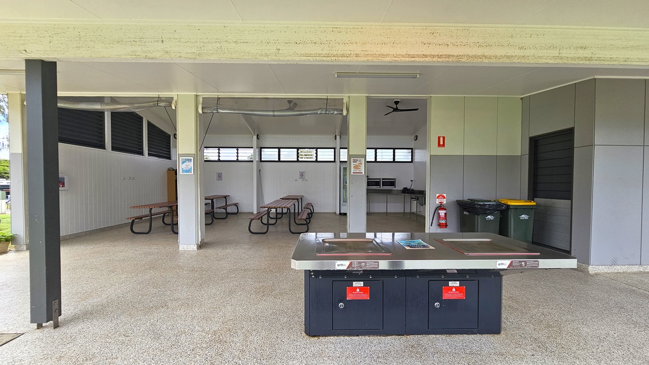 The spacious BBQ area has picnic tables and a wooden display rack in the left corner. On the right, there's a fridge, microwave ovens, and a sink. The pitched white ceiling hosts ceiling fans. Walls showcase a white timber design, with jalousy windows and light fixtures. Safety reminders are affixed to walls. In front is a barbecue grill with safety warning stickers. Two rubbish bins and a fire extinguisher are positioned on the outer wall on the right. The spacious BBQ area has picnic tables and a wooden display rack in the left corner. On the right, there's a fridge, microwave ovens, and a sink. The pitched white ceiling hosts ceiling fans. Walls showcase a white timber design, with jalousy windows and light fixtures. Safety reminders are affixed to walls. In front is a barbecue grill with safety warning stickers. Two rubbish bins and a fire extinguisher are positioned on the outer wall on the right.