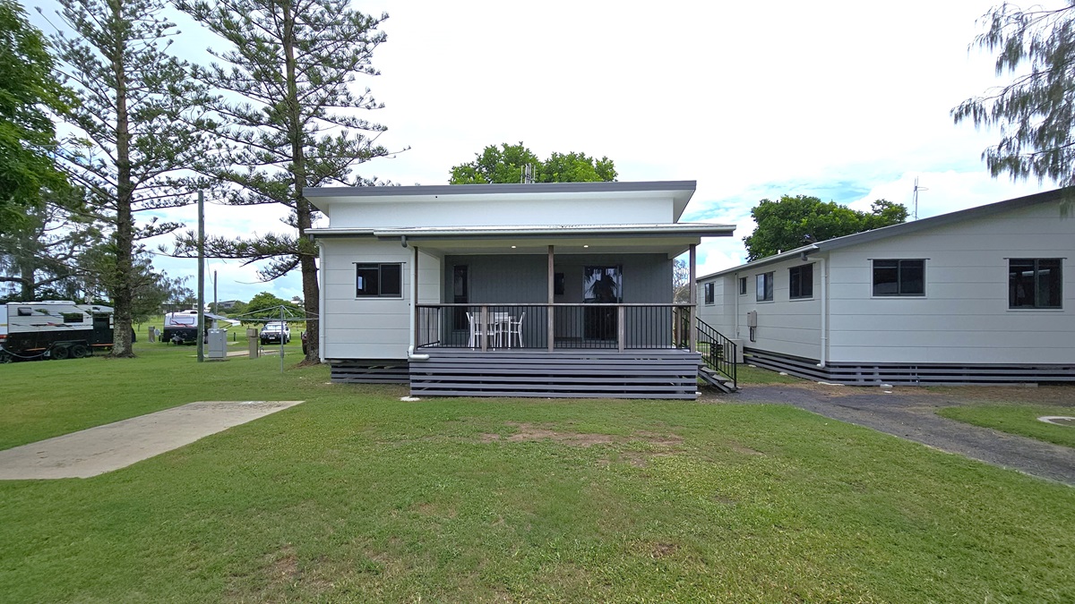 The standard cabin is a one-storey structure with a verandah, featuring steps with handrails on the right. Its exterior displays white and grey walls with pitched roofing. Tables and chairs are placed on the veranda, while another cabin stands to the right. Surrounding the area are green grass and pine trees, with trucks and vehicles visible under the shade of trees. The standard cabin is a one-storey structure with a verandah, featuring steps with handrails on the right. Its exterior displays white and grey walls with pitched roofing. Tables and chairs are placed on the veranda, while another cabin stands to the right. Surrounding the area are green grass and pine trees, with trucks and vehicles visible under the shade of trees.