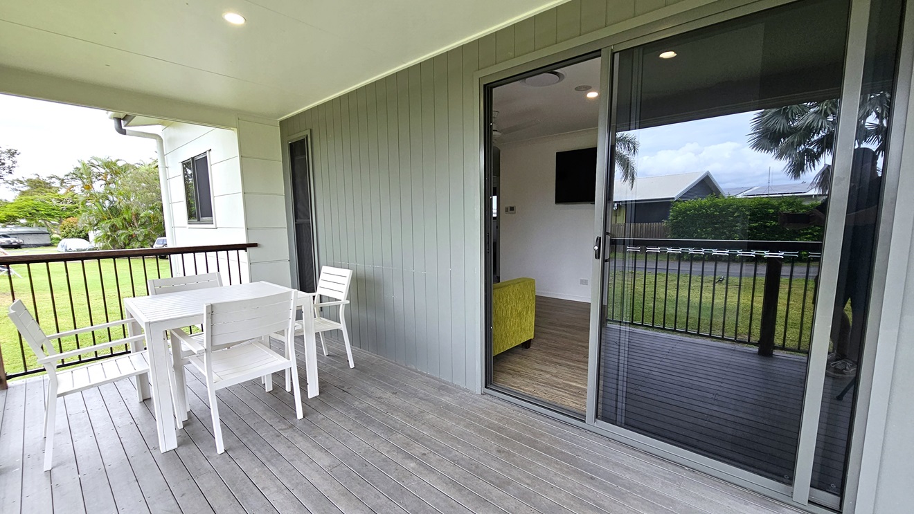 The verandah boasts a grey timber floor with white tables and chairs near metal railings. The wall exhibits a grey wooden plank design, while the ceiling is white. A glass sliding door is located to the right. Inside the cabin, a timber floor is accompanied by a green couch and a flatscreen TV mounted on a white wall. Surrounding the cabin are trees, grass, and visible vehicles. The verandah boasts a grey timber floor with white tables and chairs near metal railings. The wall exhibits a grey wooden plank design, while the ceiling is white. A glass sliding door is located to the right. Inside the cabin, a timber floor is accompanied by a green couch and a flatscreen TV mounted on a white wall. Surrounding the cabin are trees, grass, and visible vehicles.
