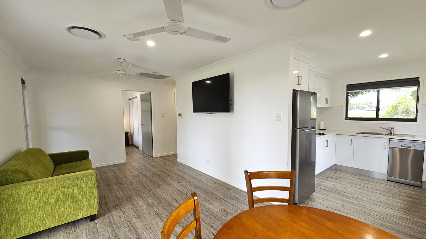 To the left is a door leading to the bedroom. Adjacent, a green couch rests against the middle wall, with a flatscreen TV mounted on the wall. To the right, a kitchen area is equipped with kitchen equipment, drawers, cabinets, and a sink. A round wooden table sits nearby. A window is positioned above the sink area. The floor is timber, the walls are white, and the ceiling features ceiling fans and LED lights. To the left is a door leading to the bedroom. Adjacent, a green couch rests against the middle wall, with a flatscreen TV mounted on the wall. To the right, a kitchen area is equipped with kitchen equipment, drawers, cabinets, and a sink. A round wooden table sits nearby. A window is positioned above the sink area. The floor is timber, the walls are white, and the ceiling features ceiling fans and LED lights.