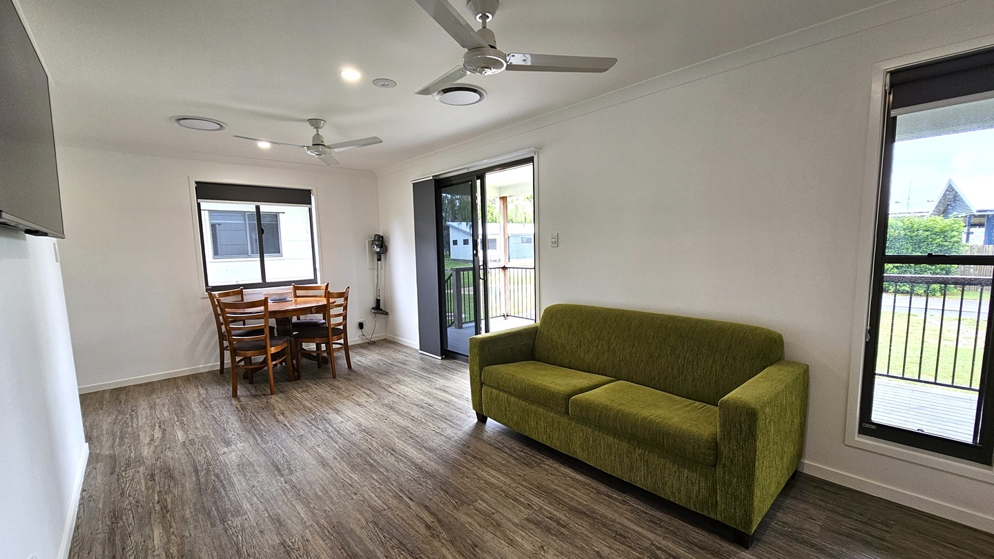 To the right is a glass sliding door leading to the verandah, alongside another door. A green couch rests against the middle wall, with a flatscreen TV mounted on it. A round wooden dining table sits near a glass window. The floor is timber, the walls are white, and the ceiling features ceiling fans and LED lights. Outside, green grass and trimmed shrubs are visible. To the right is a glass sliding door leading to the verandah, alongside another door. A green couch rests against the middle wall, with a flatscreen TV mounted on it. A round wooden dining table sits near a glass window. The floor is timber, the walls are white, and the ceiling features ceiling fans and LED lights. Outside, green grass and trimmed shrubs are visible.