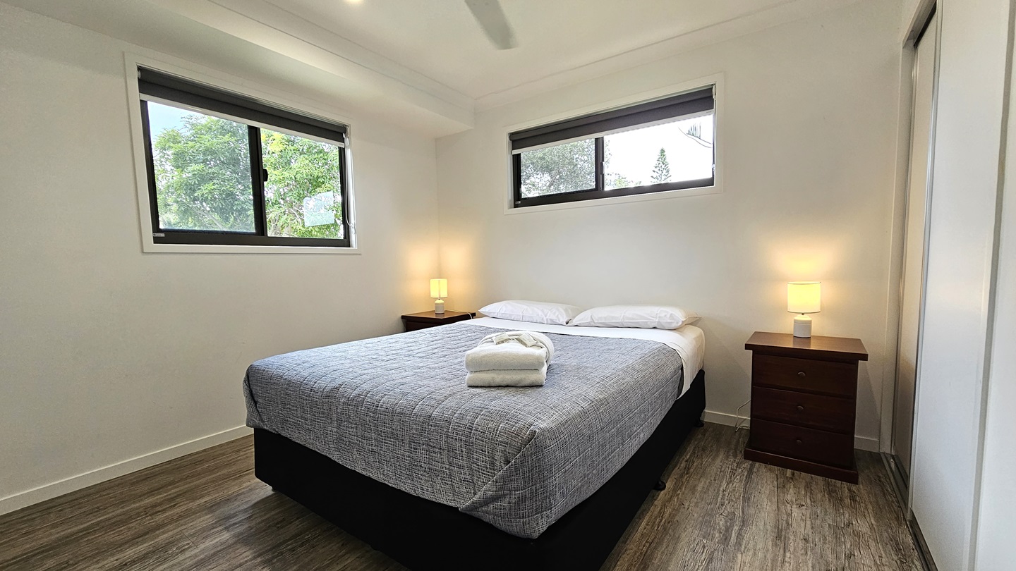In the centre stands a white queen-size bed adorned with a grey cover. White pillows and a neatly folded white towel rest atop. Flanking the bed are two bedside tables, each with a lampshade. A window is situated on the central and left walls. The walls and ceiling are painted white, illuminated by LED lights and a ceiling fan. The floor is timber. In the centre stands a white queen-size bed adorned with a grey cover. White pillows and a neatly folded white towel rest atop. Flanking the bed are two bedside tables, each with a lampshade. A window is situated on the central and left walls. The walls and ceiling are painted white, illuminated by LED lights and a ceiling fan. The floor is timber.