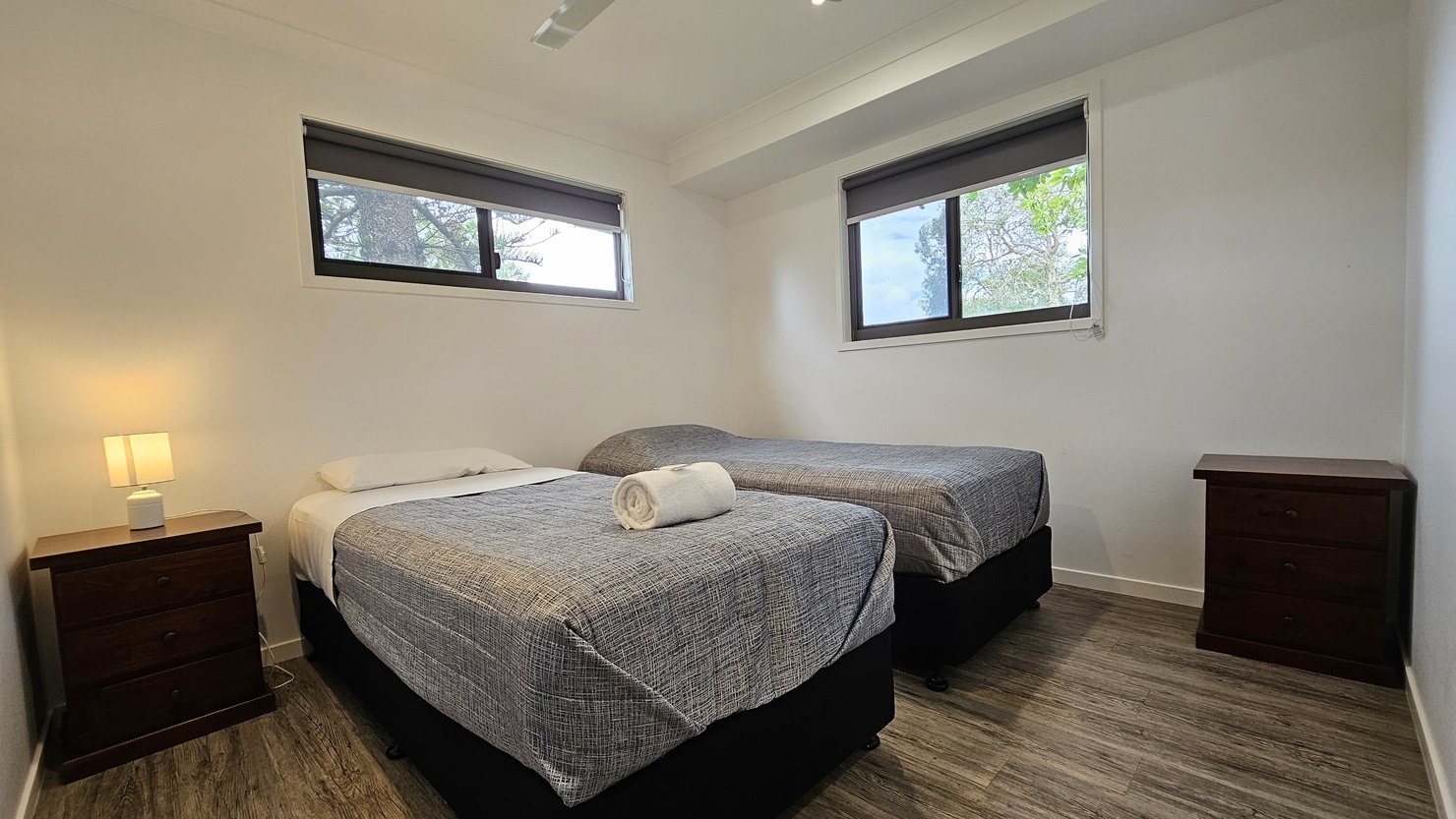 In the room, two single beds with grey covers stand opposite each other, each adorned with white pillows and a neatly rolled white towel. Windows are positioned on the middle and right walls. A bedside table with a lampshade is placed on the left corner, and another table on the right. The walls and ceiling are painted white, illuminated by LED lights and a ceiling fan. The floor is timber. In the room, two single beds with grey covers stand opposite each other, each adorned with white pillows and a neatly rolled white towel. Windows are positioned on the middle and right walls. A bedside table with a lampshade is placed on the left corner, and another table on the right. The walls and ceiling are painted white, illuminated by LED lights and a ceiling fan. The floor is timber.