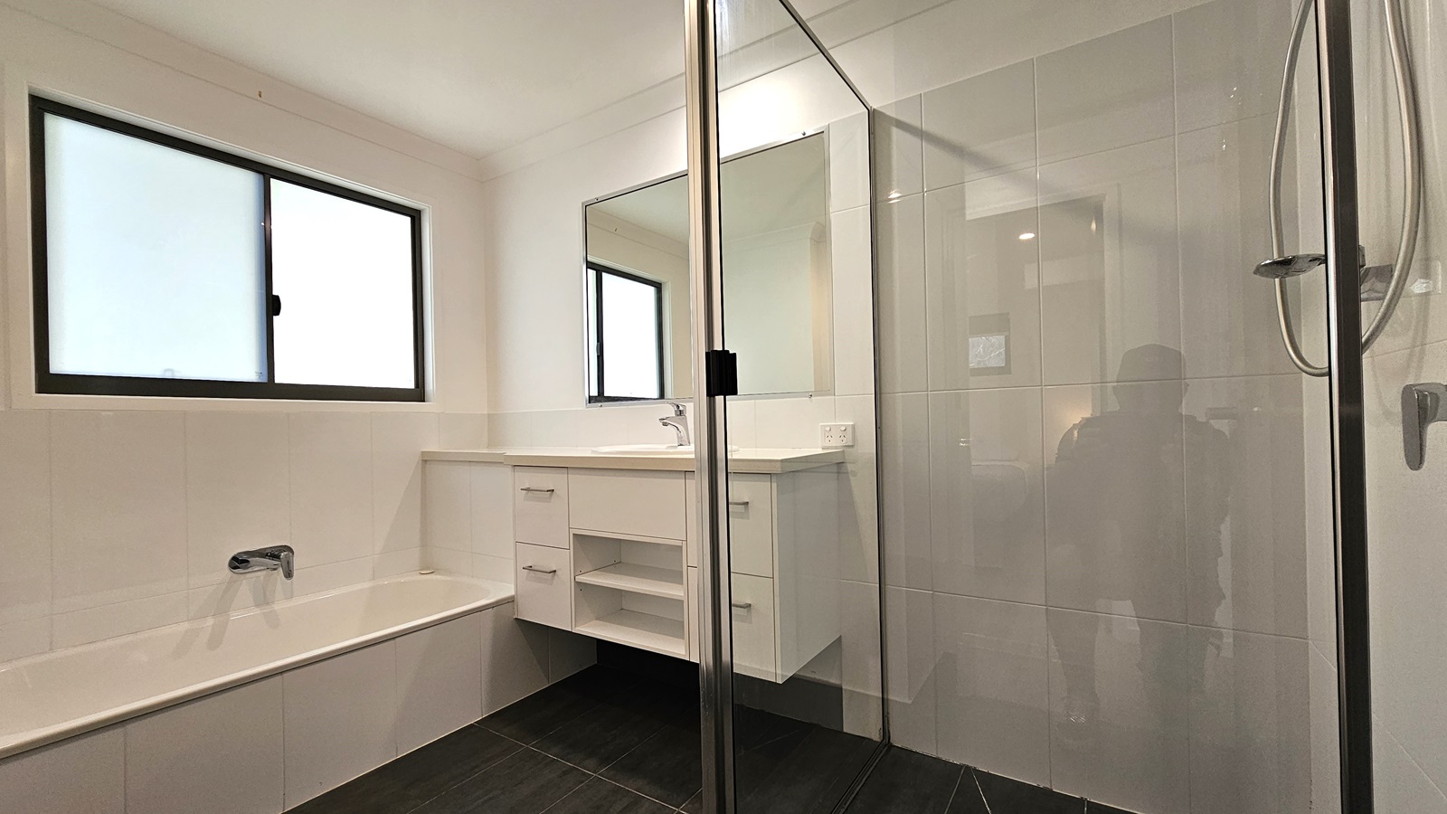 In the accessible bathroom, a shower with a handrail is positioned on the right, enclosed by a glass partition. Adjacent is a lavatory equipped with drawers and a mirror. On the left is a bathtub set against the wall, with a window positioned above it. The floor features grey tiles, while the walls and ceiling are white with LED lights. In the accessible bathroom, a shower with a handrail is positioned on the right, enclosed by a glass partition. Adjacent is a lavatory equipped with drawers and a mirror. On the left is a bathtub set against the wall, with a window positioned above it. The floor features grey tiles, while the walls and ceiling are white with LED lights.