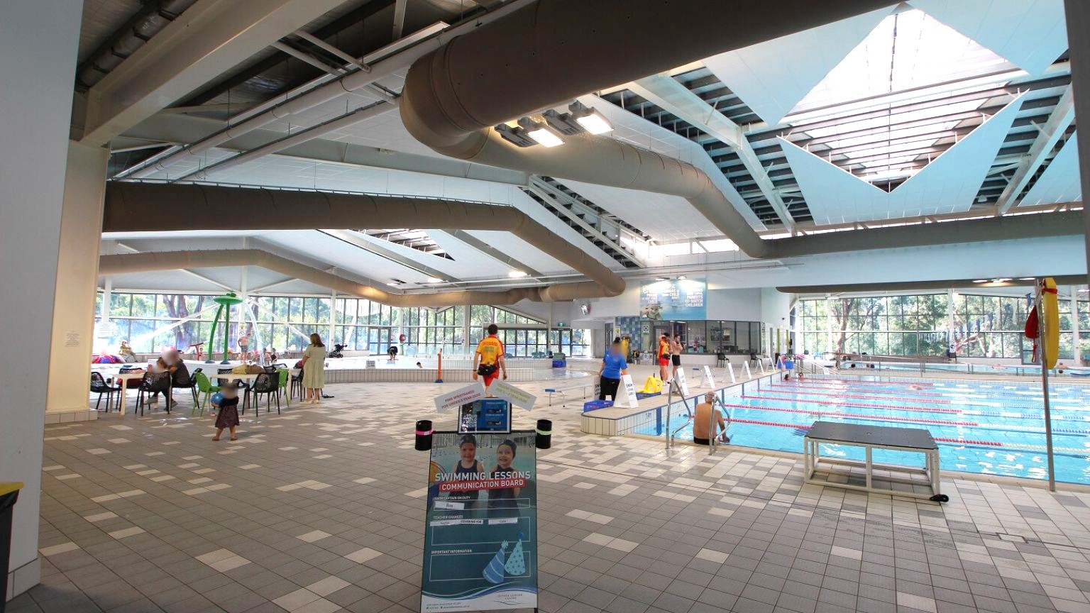 An indoor swimming pool area with a tiled floor. On the left, there are tables and chairs with people sitting and a child walking. In the centre, a sign about swimming lessons is visible. On the right, the pool has lane dividers and a few people standing by the edge. The ceiling features large, geometric skylights, and the background shows large windows with a view of trees outside. An indoor swimming pool area with a tiled floor. On the left, there are tables and chairs with people sitting and a child walking. In the centre, a sign about swimming lessons is visible. On the right, the pool has lane dividers and a few people standing by the edge. The ceiling features large, geometric skylights, and the background shows large windows with a view of trees outside.