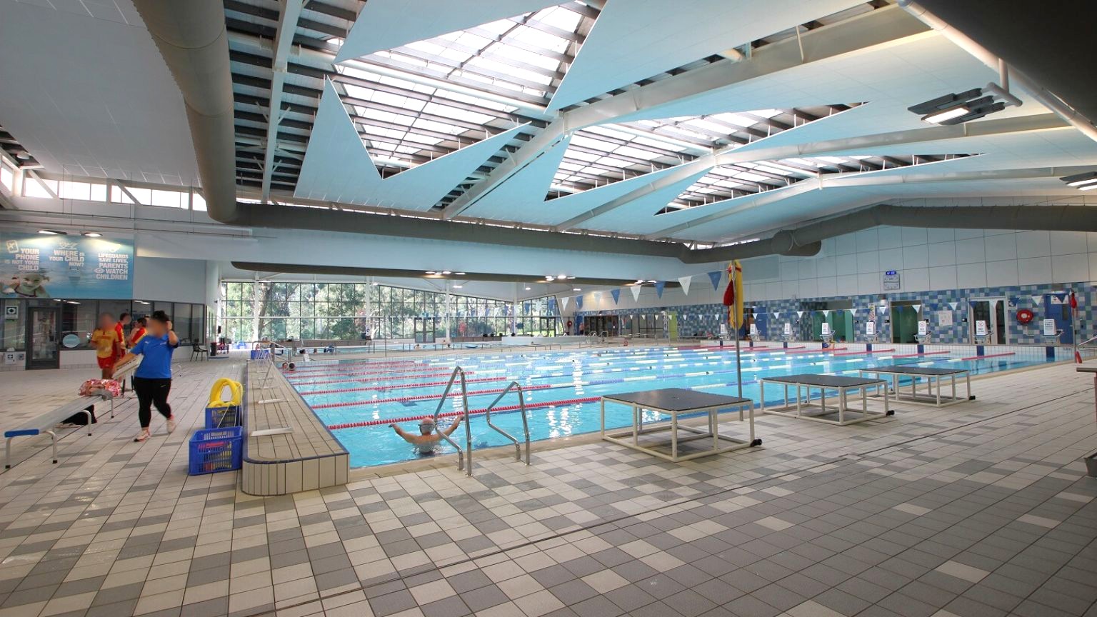 An indoor swimming pool with multiple lanes, surrounded by a tiled deck. On the left, a few people are walking near the poolside, with some equipment like a yellow lifebuoy and a blue crate nearby. The ceiling features large, angular skylights allowing natural light to illuminate the space. The walls have large windows and some signage. The floor is covered with multicoloured tiles, and there are diving platforms on the right side of the pool. An indoor swimming pool with multiple lanes, surrounded by a tiled deck. On the left, a few people are walking near the poolside, with some equipment like a yellow lifebuoy and a blue crate nearby. The ceiling features large, angular skylights allowing natural light to illuminate the space. The walls have large windows and some signage. The floor is covered with multicoloured tiles, and there are diving platforms on the right side of the pool.