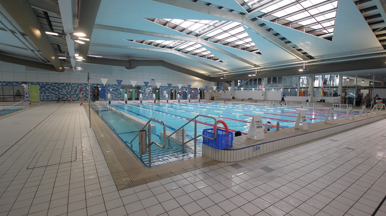 Indoor swimming pool with multiple lanes, surrounded by a tiled deck. On the left, there's a wide walkway with a non-slip surface. The right side features a row of windows and seating areas. The ceiling has large skylights allowing natural light to illuminate the space. Safety signs and pool equipment are visible around the pool's edge. Indoor swimming pool with multiple lanes, surrounded by a tiled deck. On the left, there's a wide walkway with a non-slip surface. The right side features a row of windows and seating areas. The ceiling has large skylights allowing natural light to illuminate the space. Safety signs and pool equipment are visible around the pool's edge.