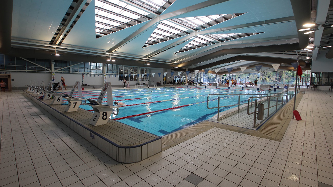 Indoor swimming pool with multiple lanes, each marked with numbers on starting blocks. The pool is surrounded by a tiled deck. The ceiling features large skylights and angular beams, allowing natural light to illuminate the area. On the right, there are handrails leading into the pool, and a few people are visible swimming and standing by the poolside. The background shows large windows and some seating areas. Indoor swimming pool with multiple lanes, each marked with numbers on starting blocks. The pool is surrounded by a tiled deck. The ceiling features large skylights and angular beams, allowing natural light to illuminate the area. On the right, there are handrails leading into the pool, and a few people are visible swimming and standing by the poolside. The background shows large windows and some seating areas.